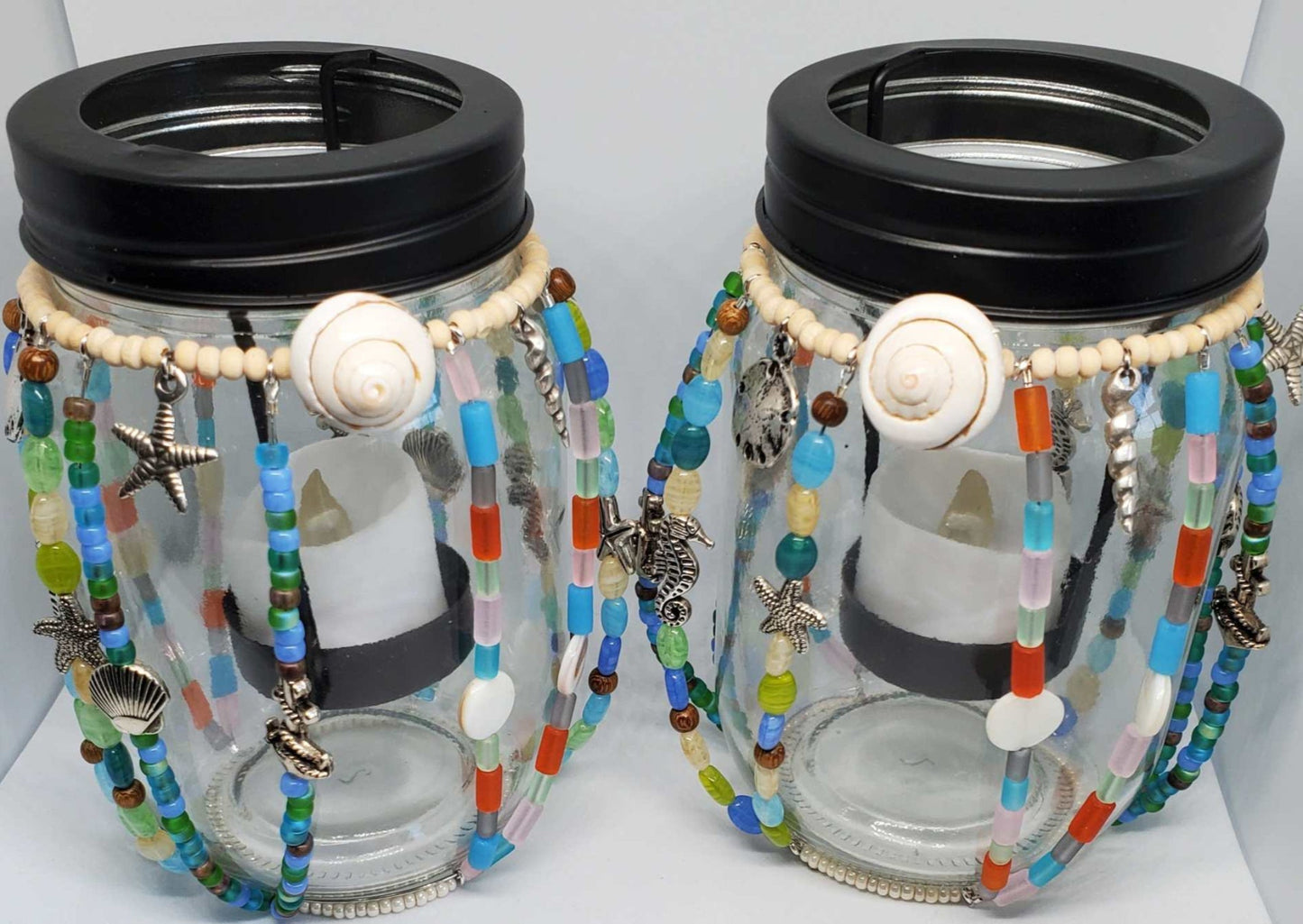 Pair of Mason jar candle holders decorated with colorful beads and ocean-themed pewter charms including shells and starfish with black lids and battery-operated tealight candles inside