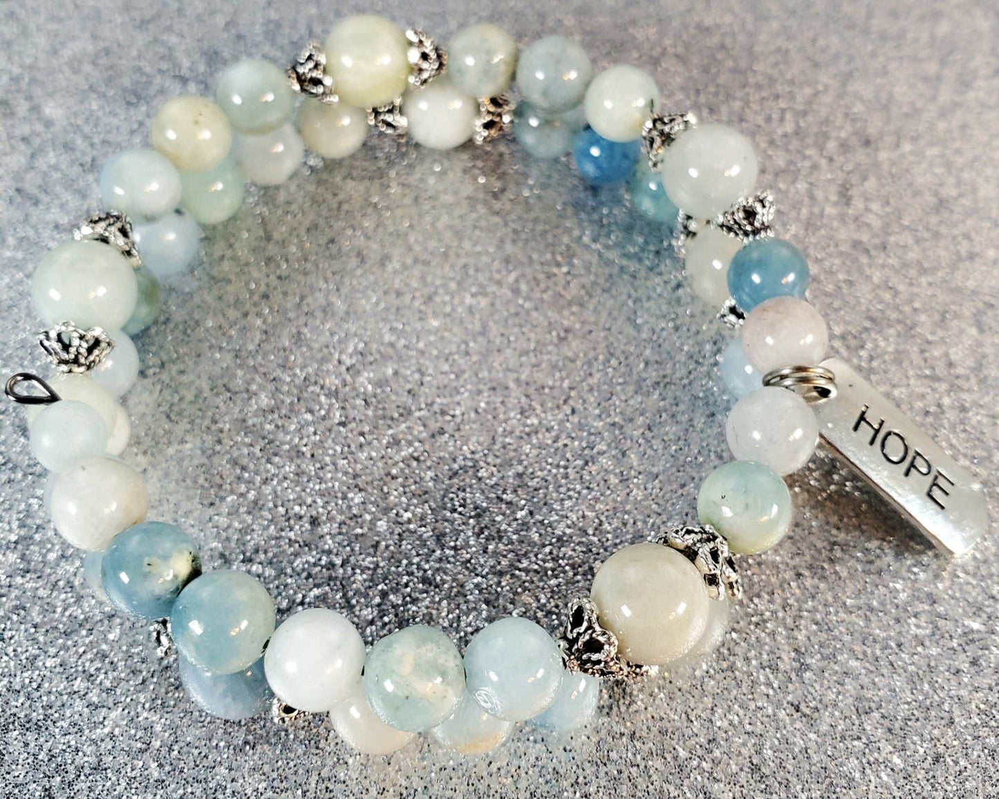 Adjustable gemstone bracelet with light blue and white beads and a pewter charm engraved with the word "Hope" on a sparkly silver background