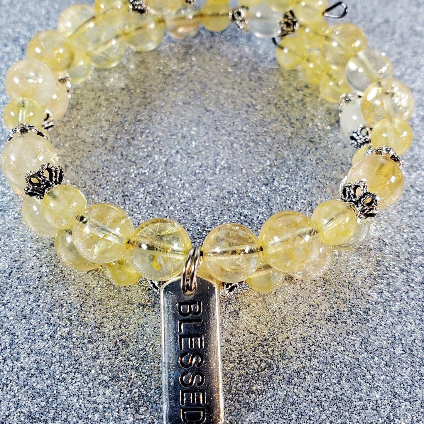 Yellow citrine gemstone bracelet with silver "Blessed" charm and decorative bead accents on a glittery surface