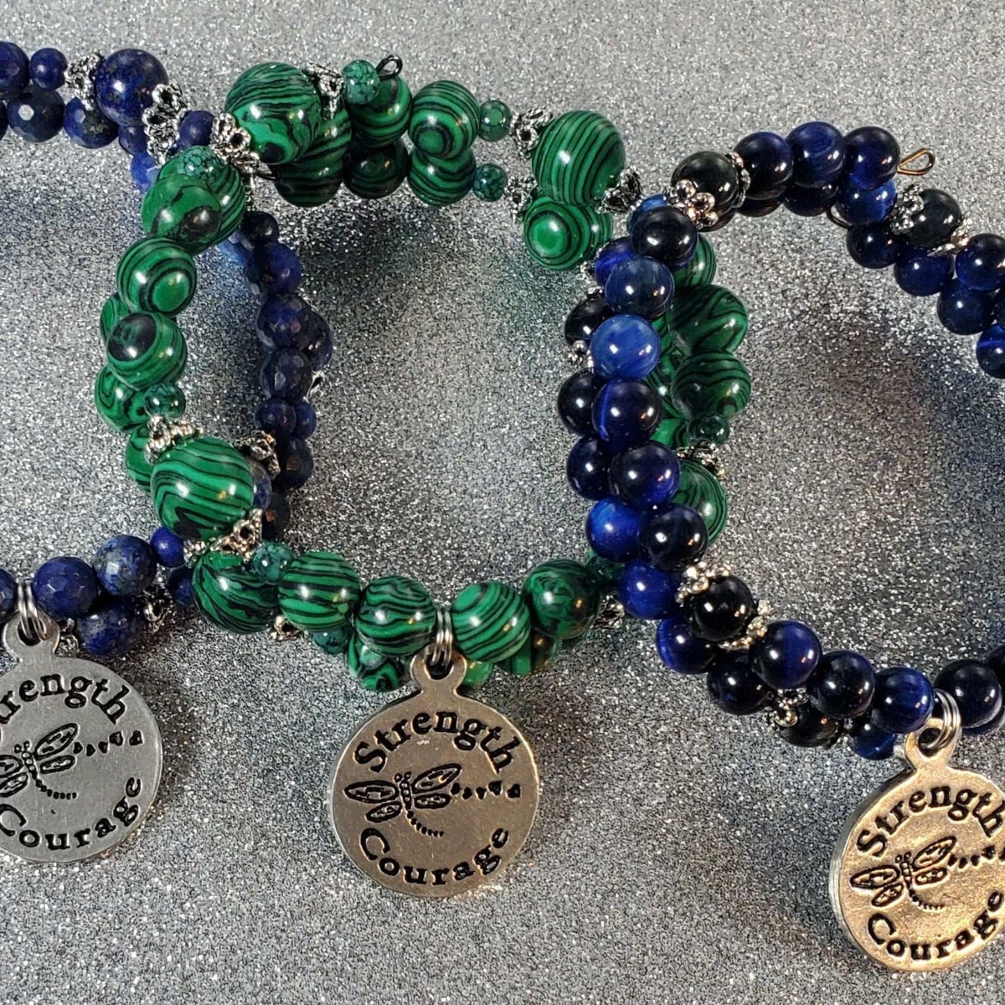 Close-up of handcrafted gemstone bracelets with green malachite and blue lapis lazuli beads featuring a "Strength & Courage" pewter charm on flexible memory wire bands.