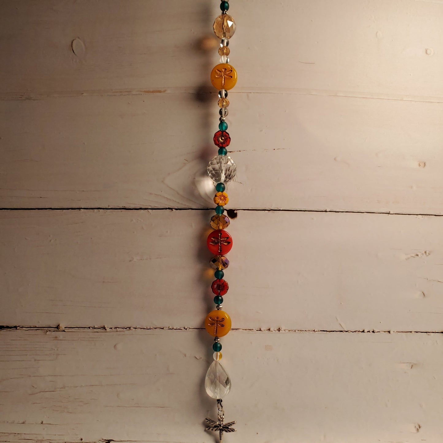 A beaded hanging suncatcher with colorful round and faceted beads, including orange, red, teal, and clear glass, suspended against a rustic white-painted wooden plank background.