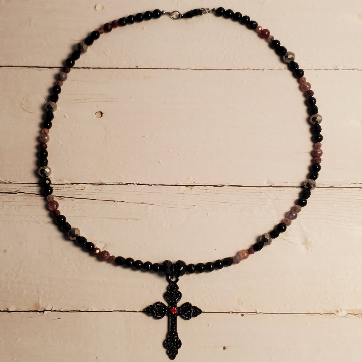 A beaded necklace with a cross pendant, featuring dark beads with subtle pinkish-red accents, displayed against a rustic white wooden background.
