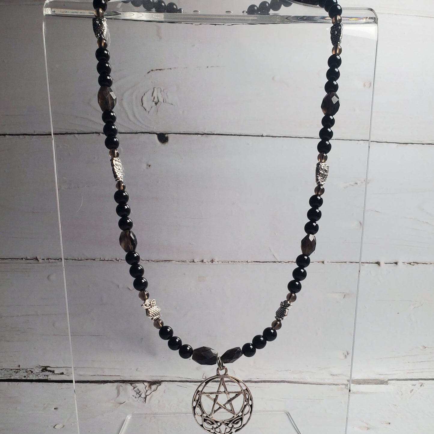 A beaded necklace with black and dark gray beads, silver-toned spacer beads, and a circular pentagram pendant, displayed on a clear acrylic stand against a white wood plank background.