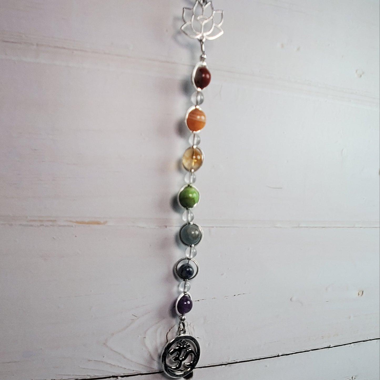 A vertical chakra bead hanging ornament with silver-toned metal links, colorful gemstone beads, a lotus flower at the top, and a circular Om symbol charm at the bottom, displayed against a white wooden wall.