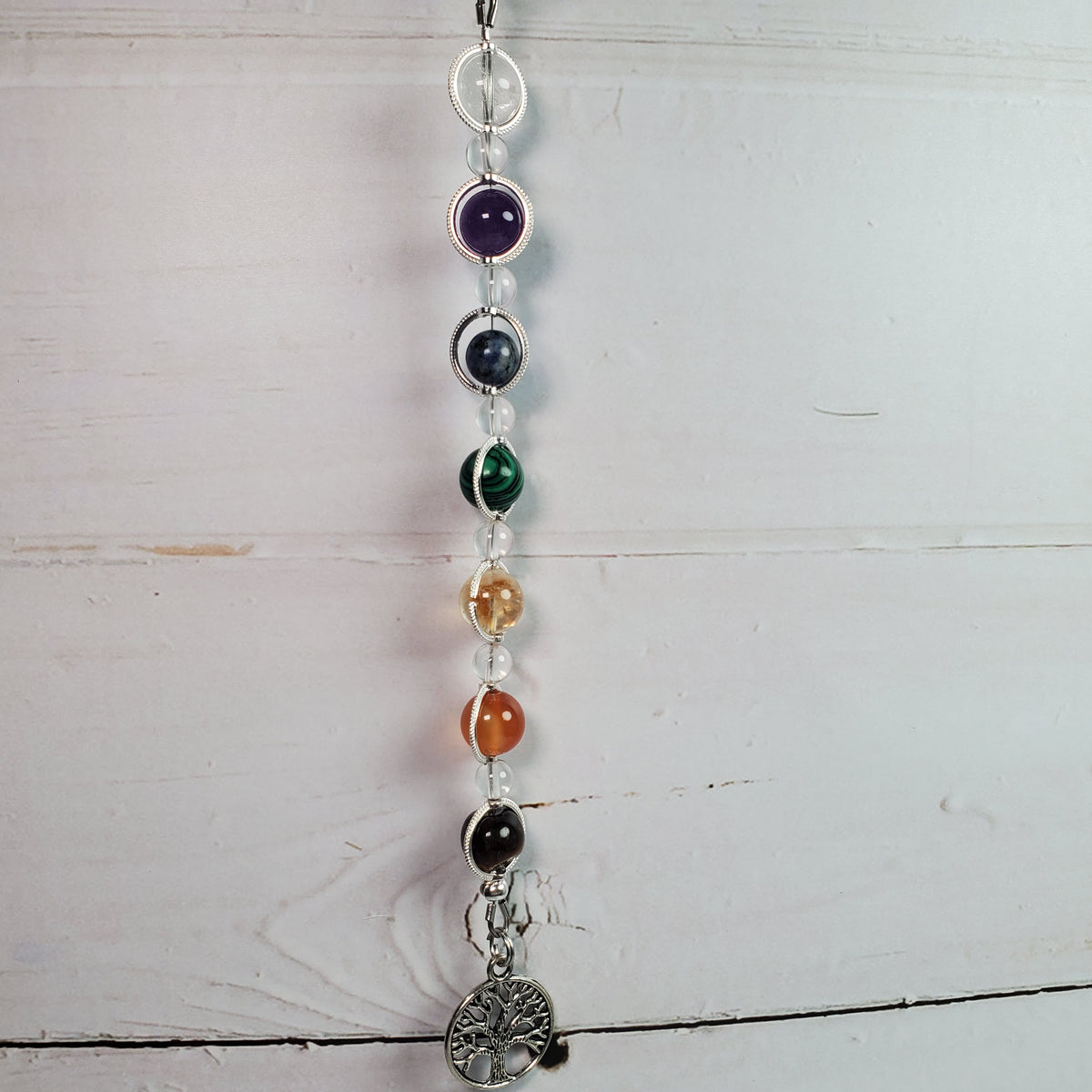 A vertical bead garland with seven colored gemstone beads and a silver tree of life pendant, hanging against a white wooden background.