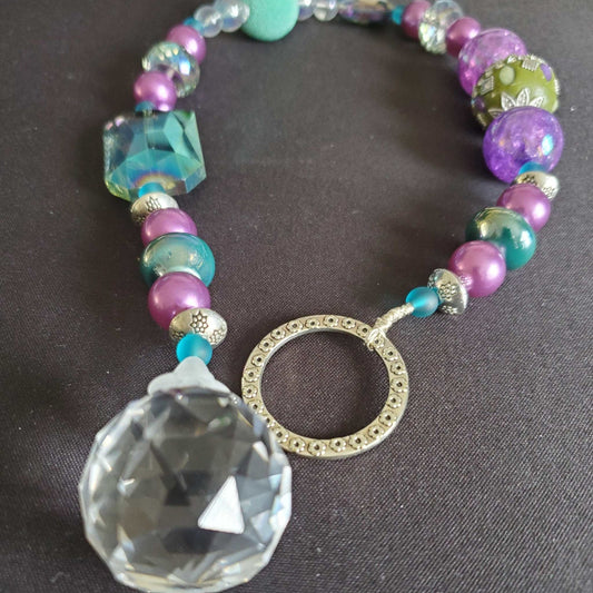 Close-up of a colorful crystal suncatcher featuring purple, green, and turquoise beads with a large faceted prism crystal pendant on a dark background