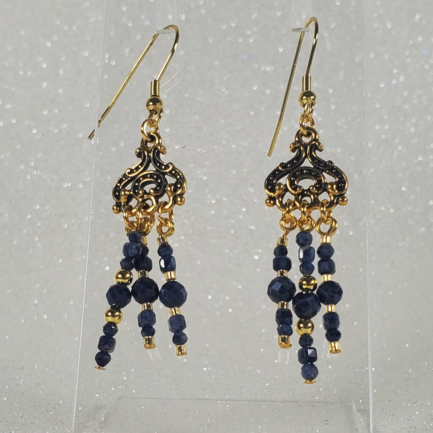 Pair of gold and blue beaded earrings on a clear stand against a silver glittery background
