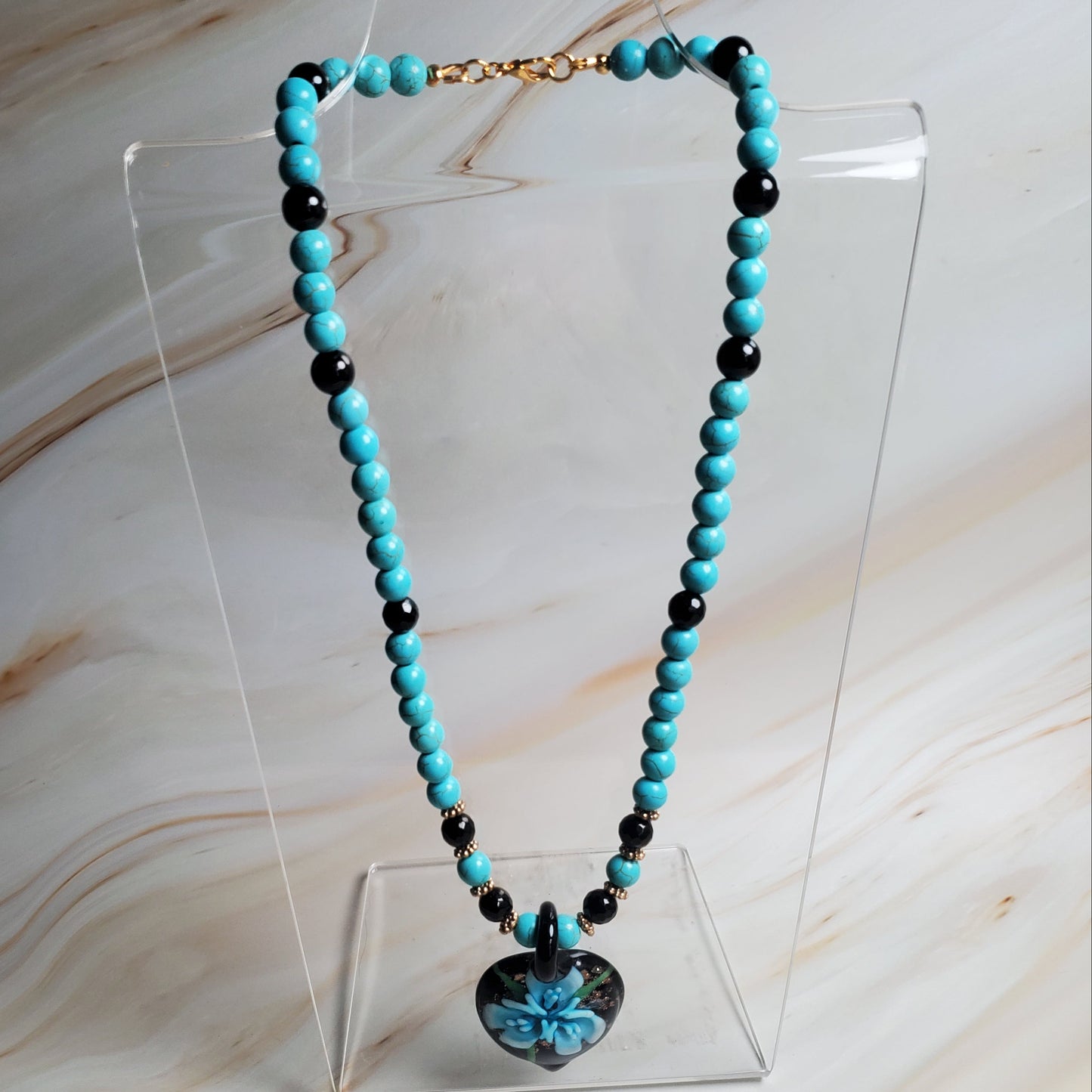 Turquoise and black beaded necklace on a clear stand against a marble background
