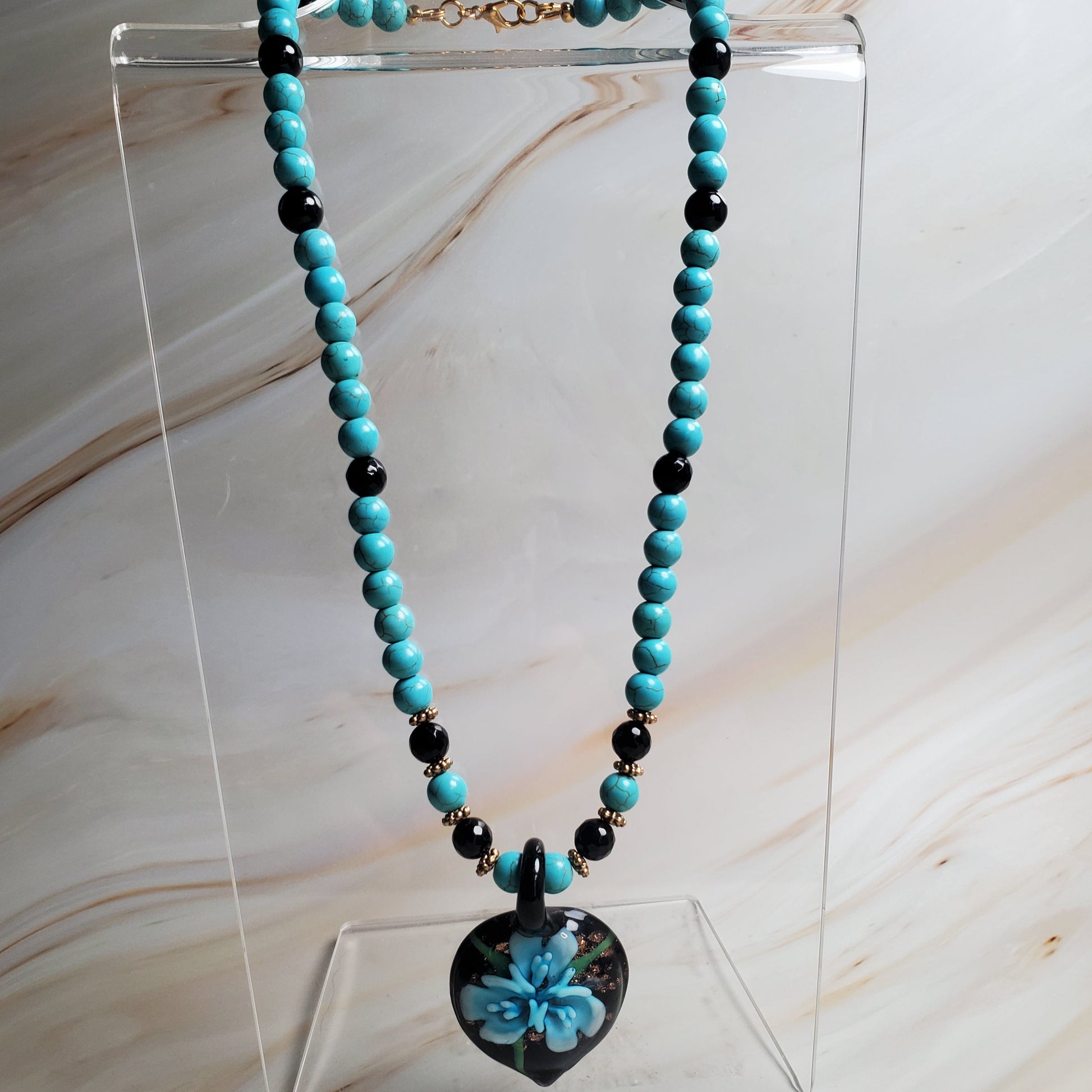 Turquoise beaded necklace with a black and blue floral pendant on a clear stand against a marble background.