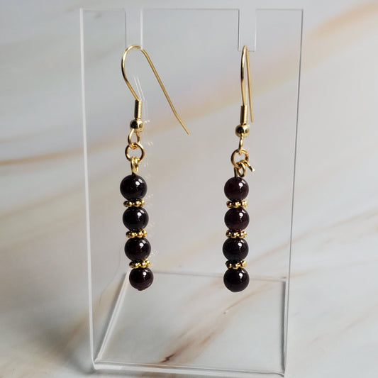 Pair of gold earrings with Garnet beads on a clear stand against a marble background