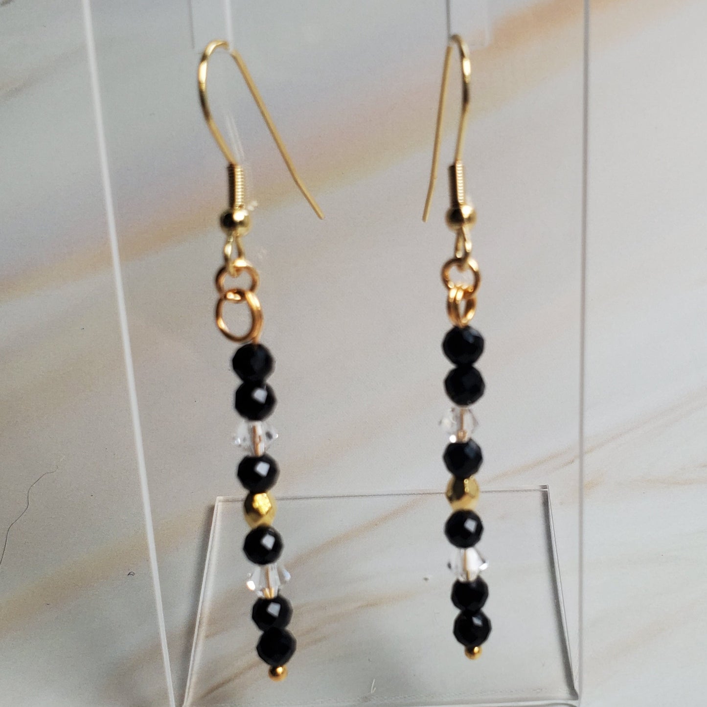 Pair of gold and black beaded earrings on a clear stand against a marble background