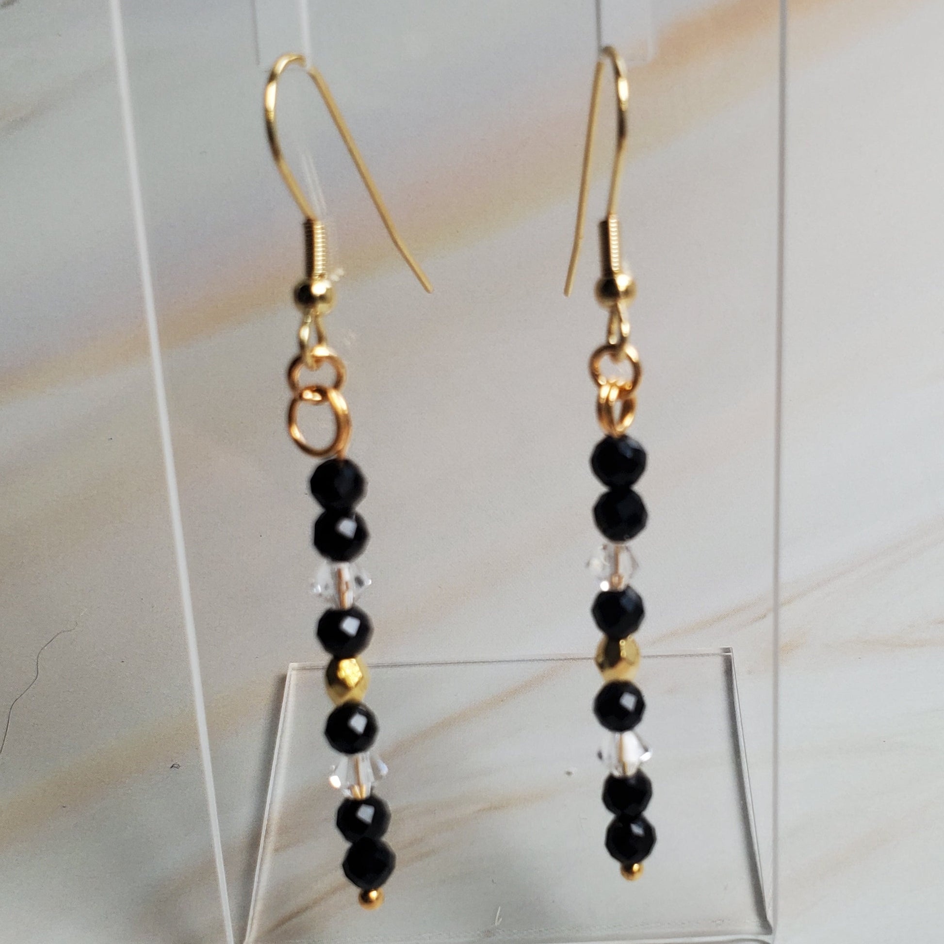 Pair of gold and black beaded earrings on a clear stand against a marble background