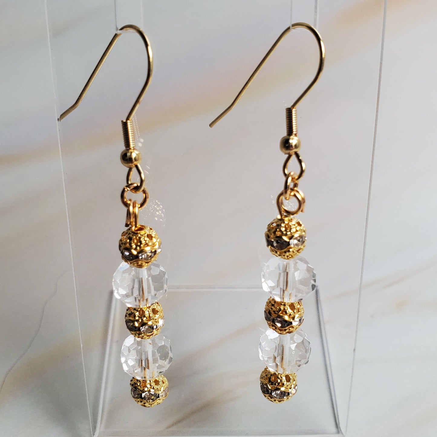 Gold and clear beaded earrings on a clear stand against a marble background