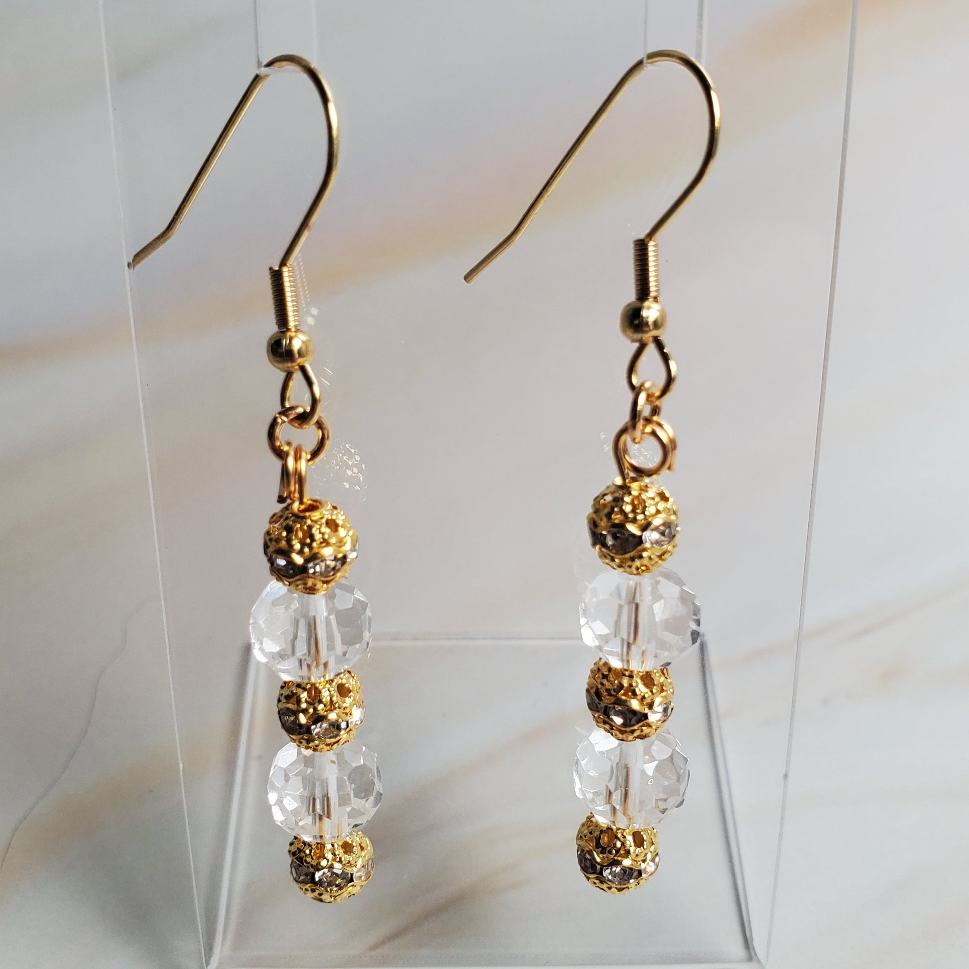 Gold and clear beaded earrings on a clear stand against a marble background