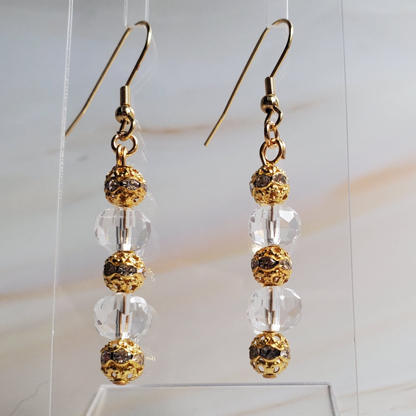Gold and clear beaded earrings on a clear stand with a marble background