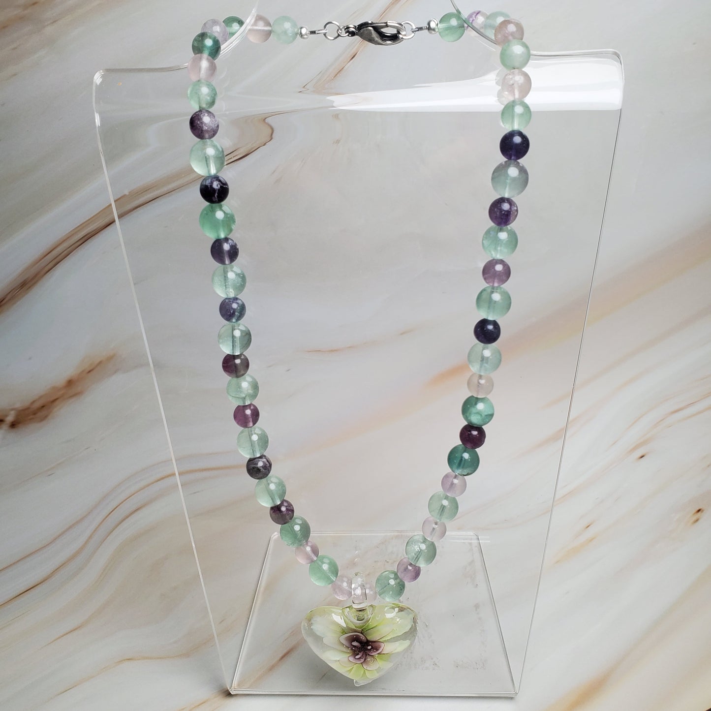 Necklace with multicolored beads and a floral clasp on a clear stand against a marble background