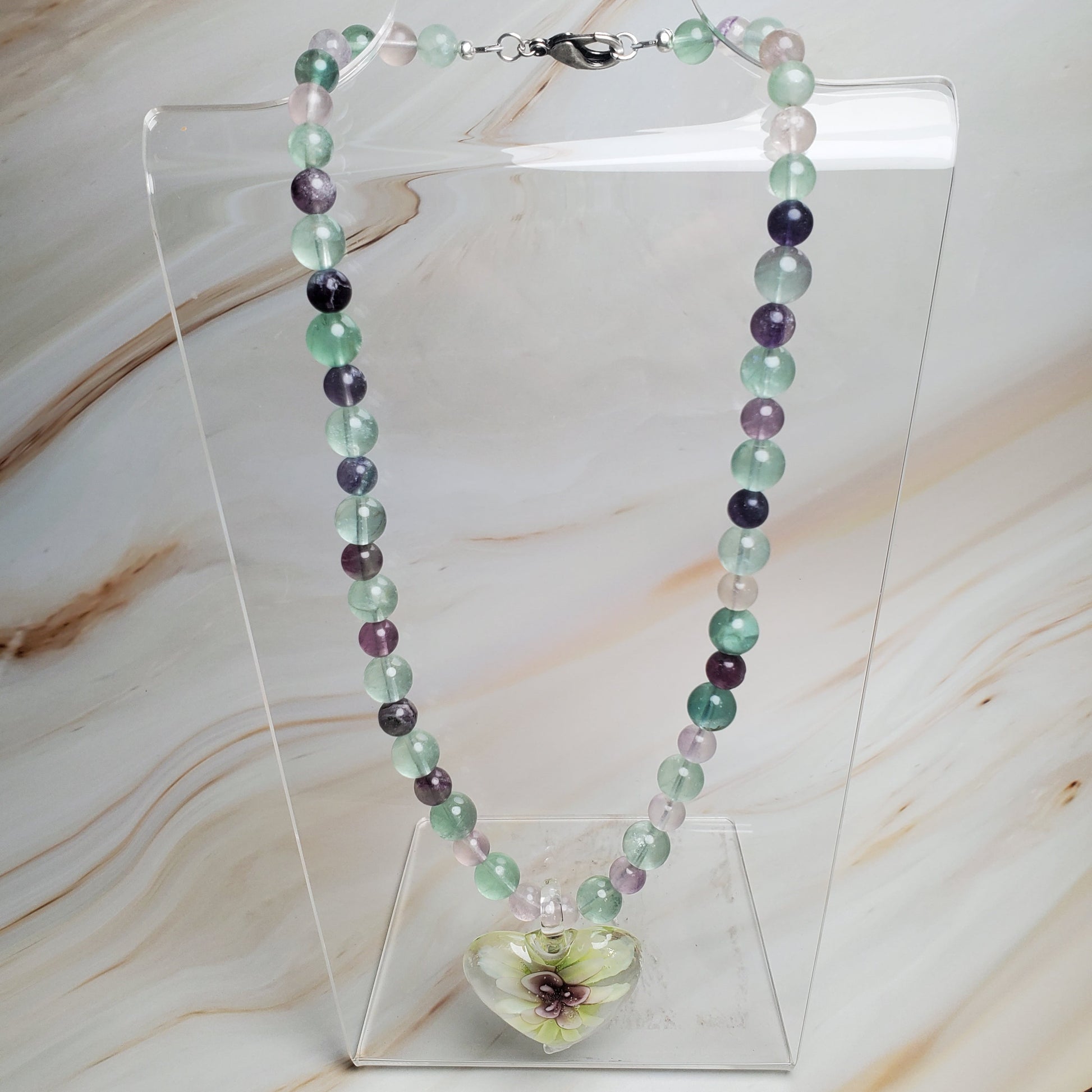 Necklace with multicolored beads and a floral clasp on a clear stand against a marble background