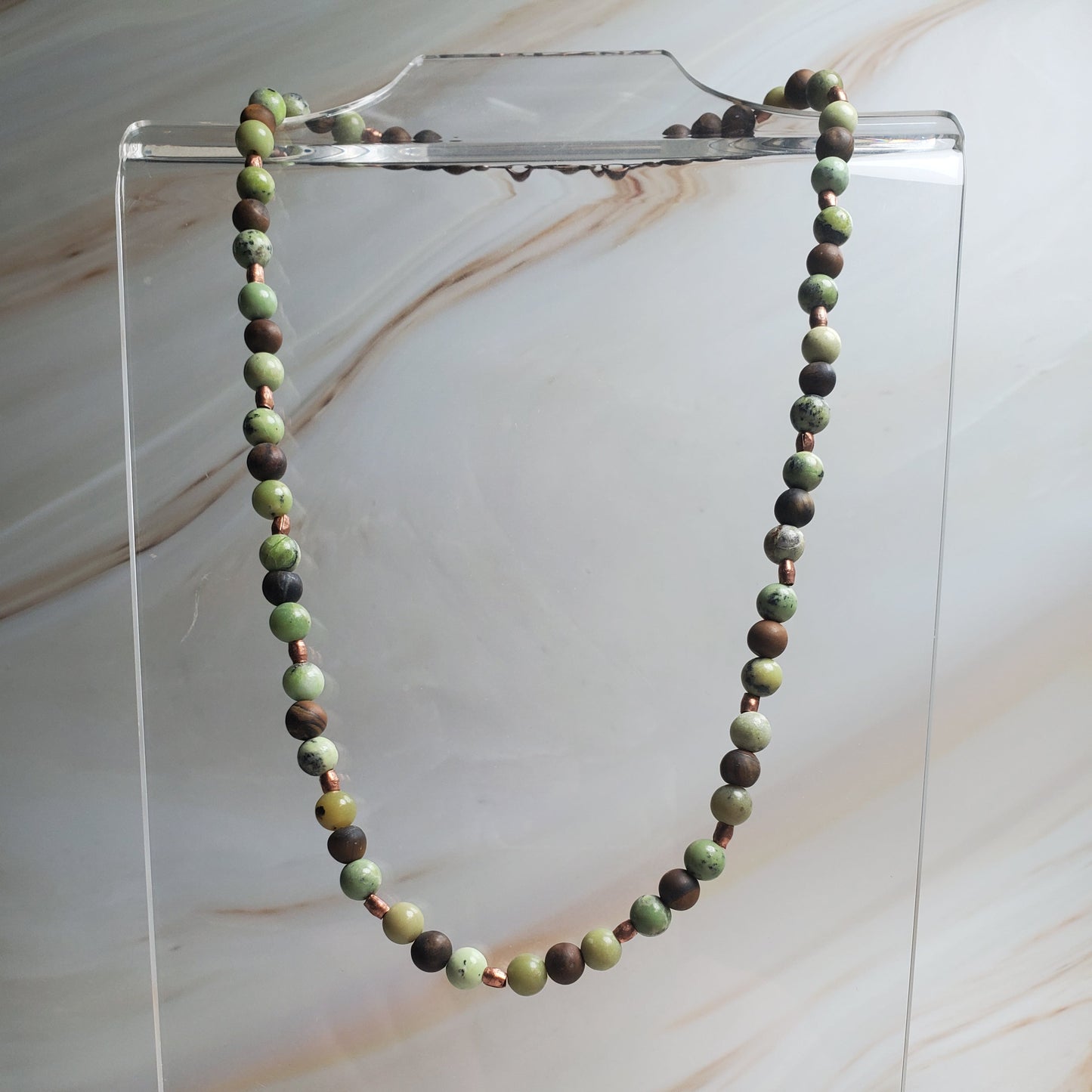 Necklace with green and brown beads on a clear stand against a marble background
