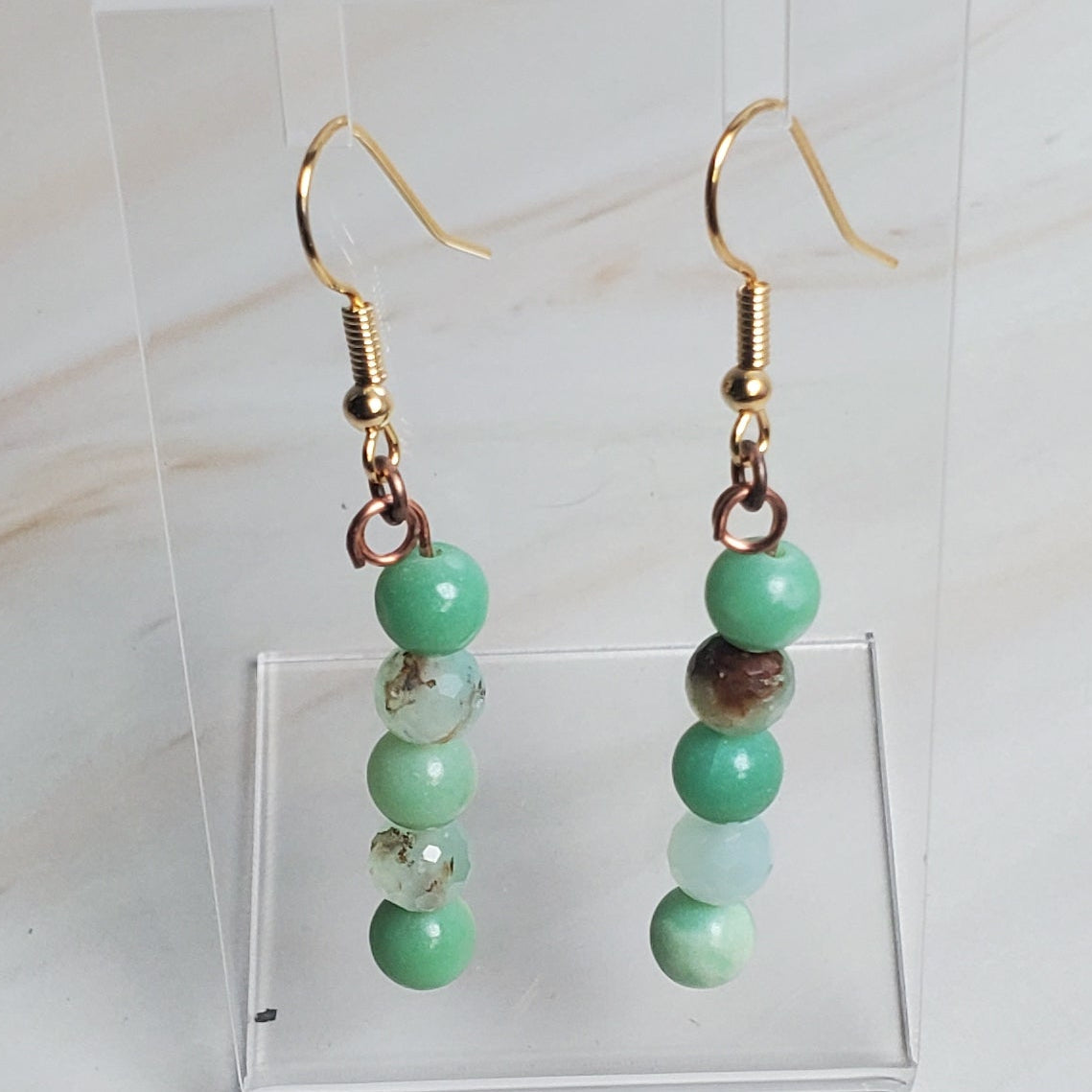 Pair of earrings with green beads on a clear stand against a marble background