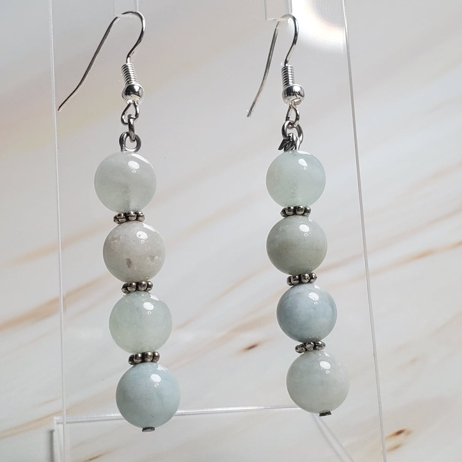Pair of earrings with light blue beads on a clear stand against a marble background