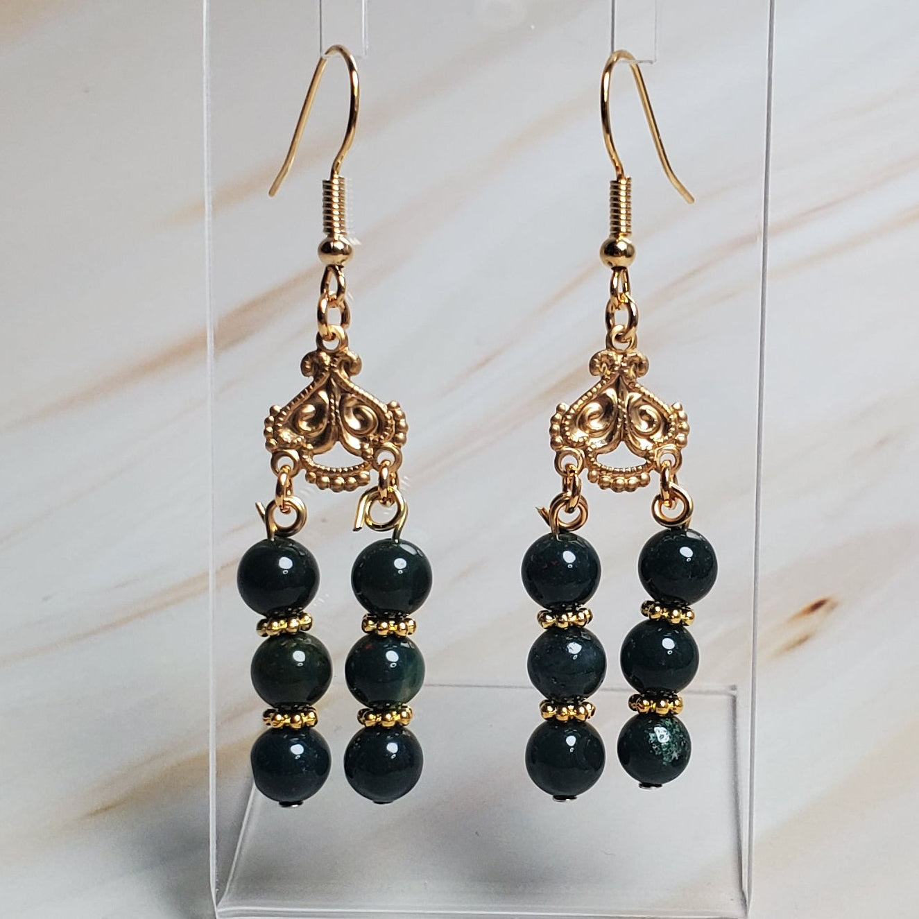 Dark Green beaded earrings on a clear stand against a marble background