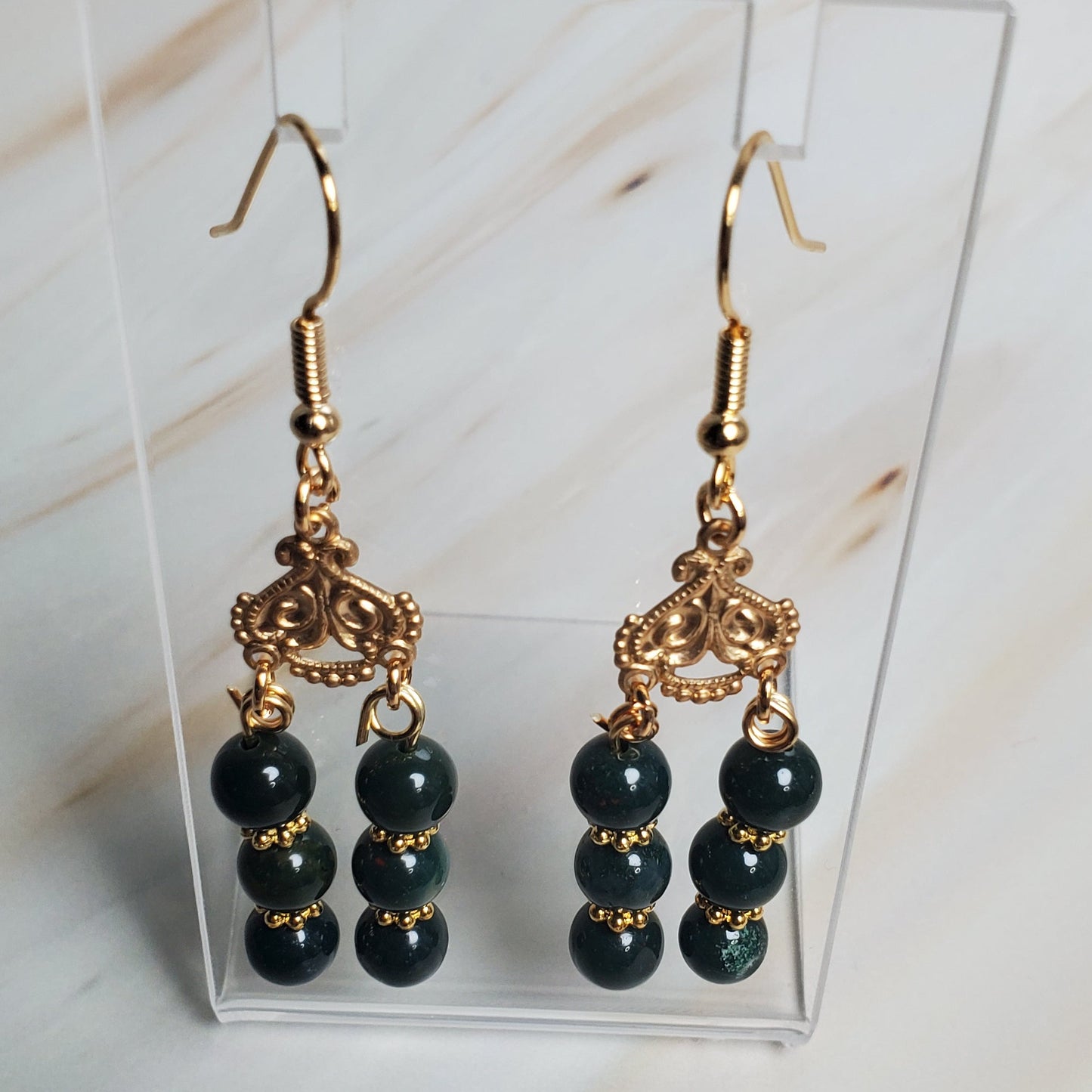 Pair of dark Green beaded earrings with gold hooks on a clear display card against a marble background.
