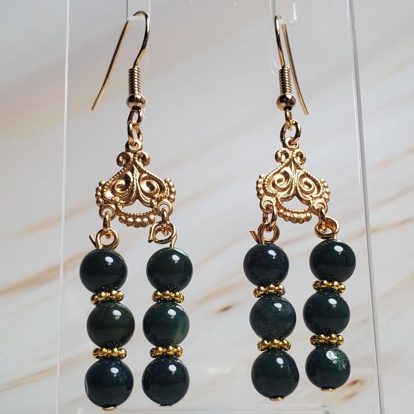 Dark Green beaded earrings with gold accents on a clear stand against a marble background