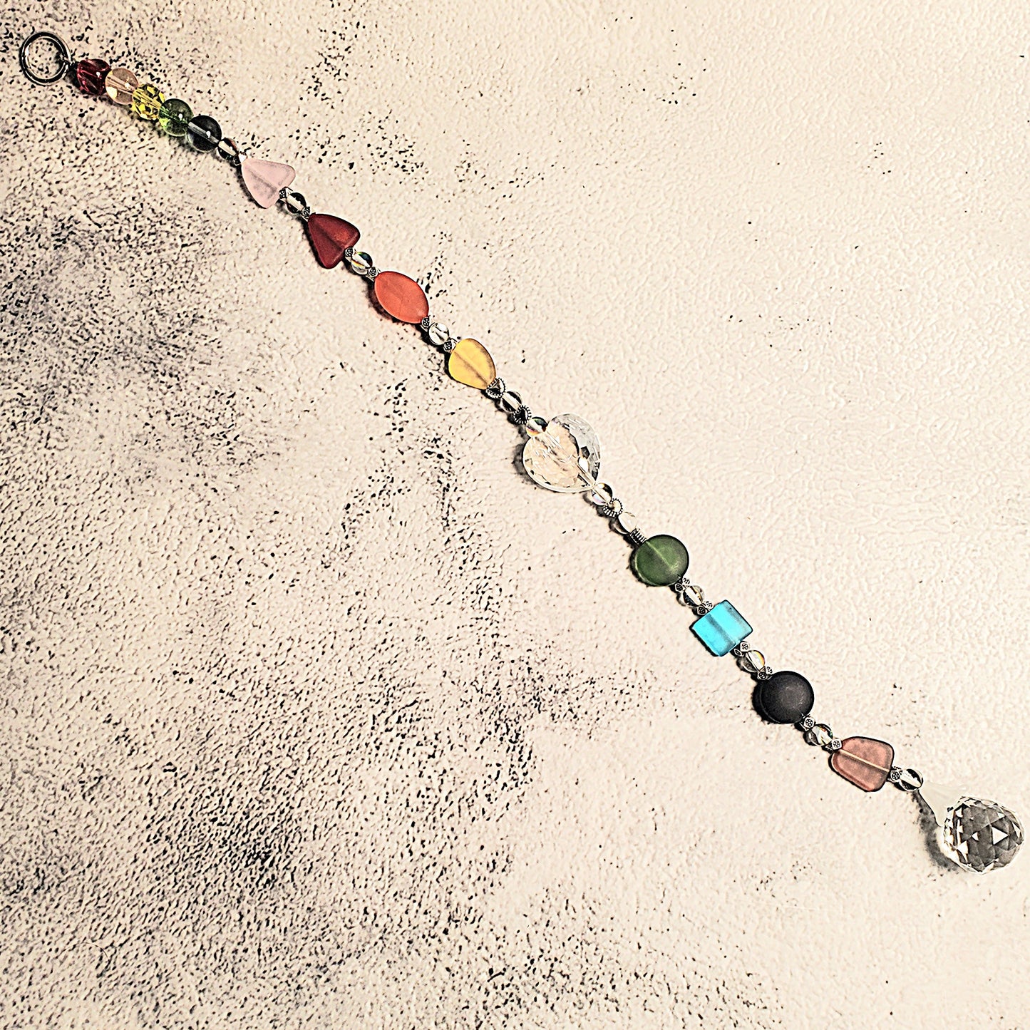 A handmade beaded Suncatcher with colorful, irregularly shaped glass beads in red, orange, yellow, green, blue, purple, and pink, connected by silver-toned chains. The suncatcher is laid out diagonally on a textured, off-white surface.