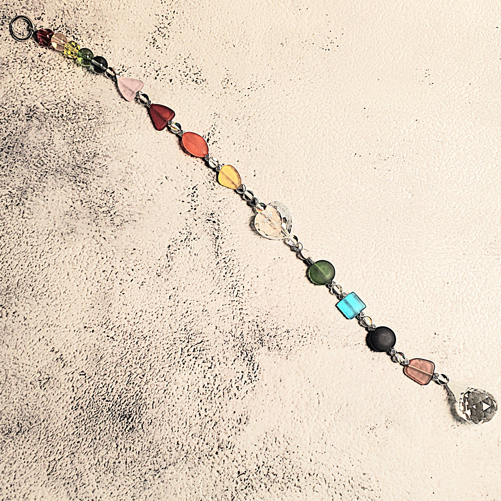 A handmade beaded Suncatcher with colorful, irregularly shaped glass beads in red, orange, yellow, green, blue, purple, and pink, connected by silver-toned chains. The suncatcher is laid out diagonally on a textured, off-white surface.