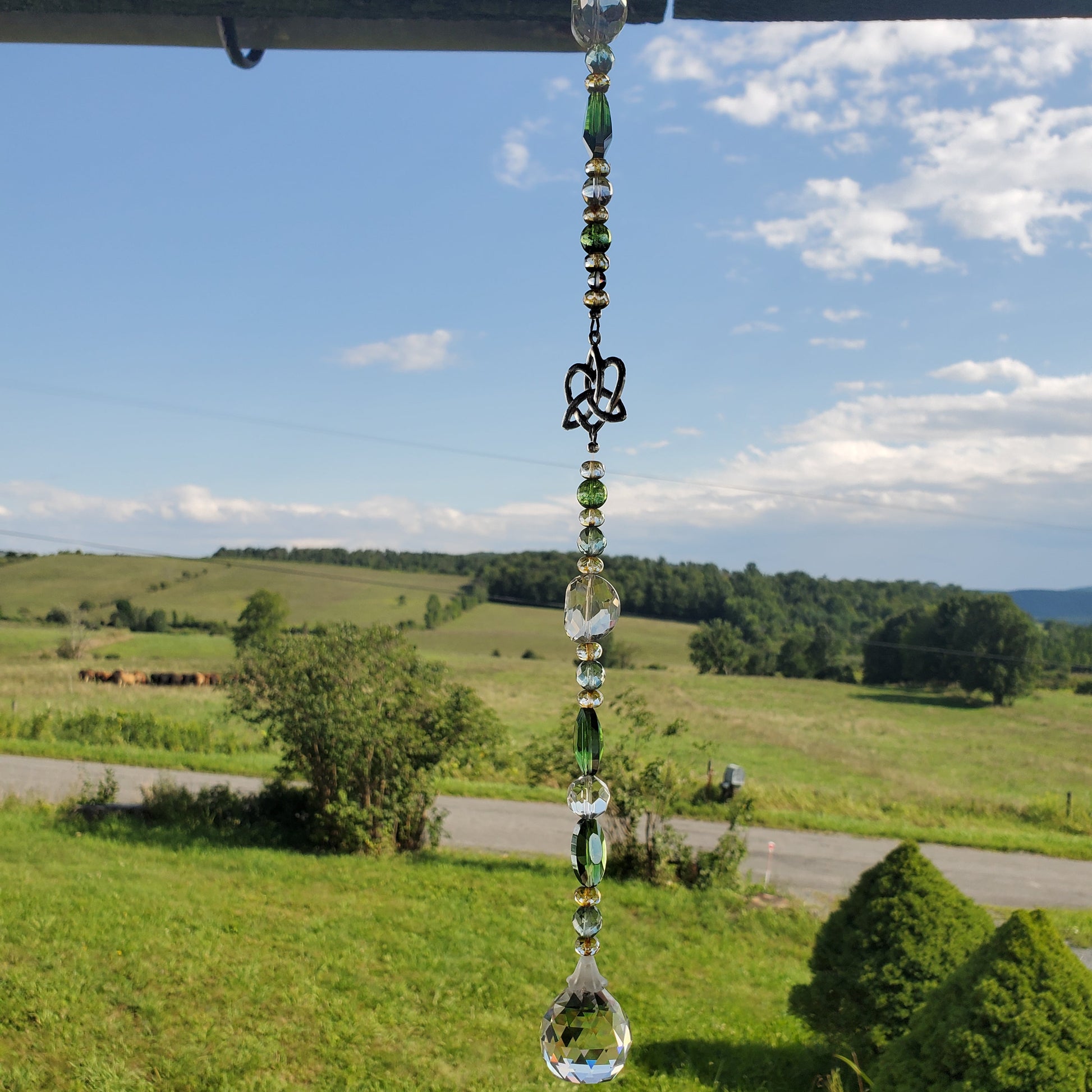 A hanging clear glass suncatcher with green beads and a decorative metal charm, suspended outdoors against a scenic background of green hills, trees, and a blue sky with clouds.