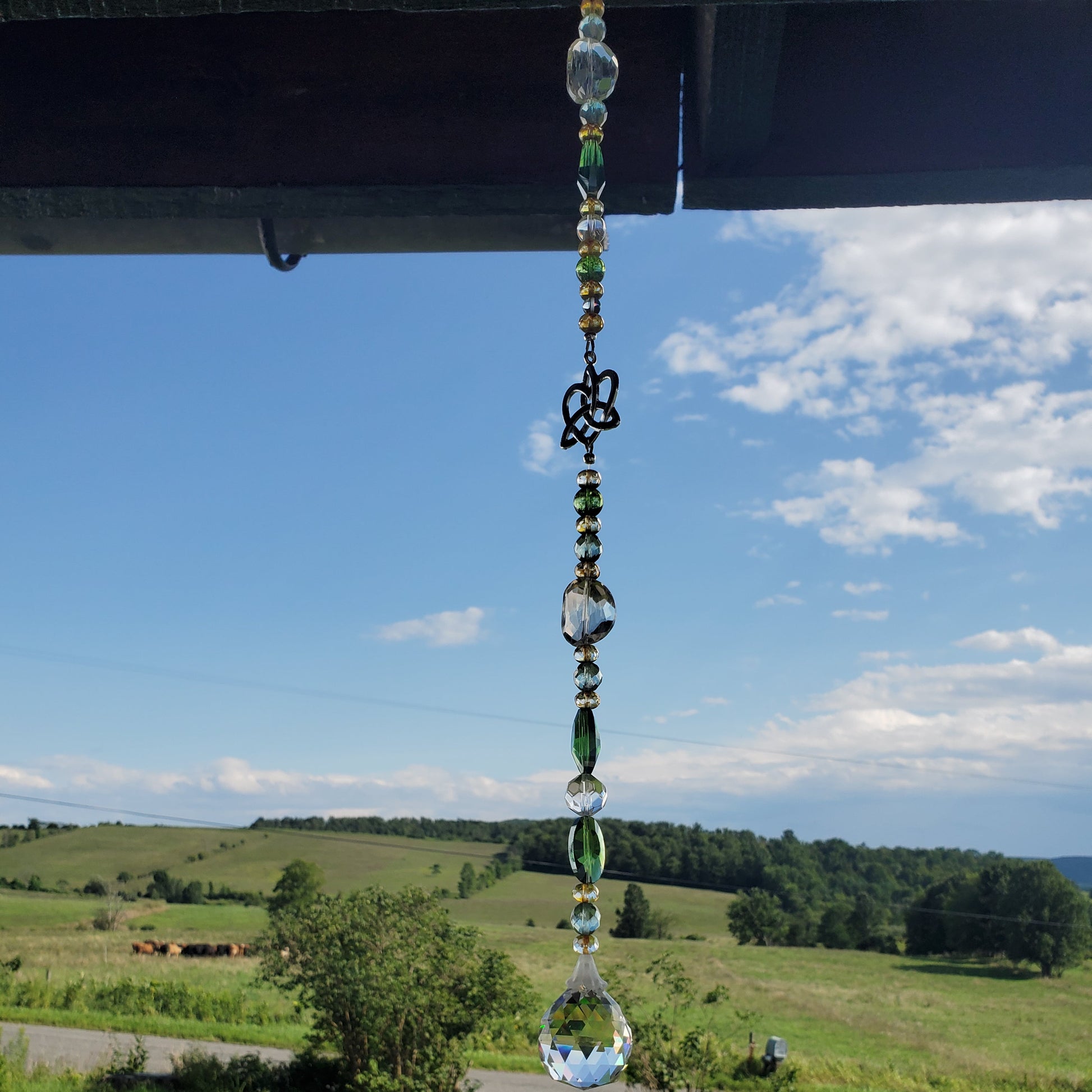 A beaded crystal suncatcher with a Celtic knot heart and a teardrop-shaped clear glass end, hanging outdoors under a roofline with a scenic green field and blue sky in the background.