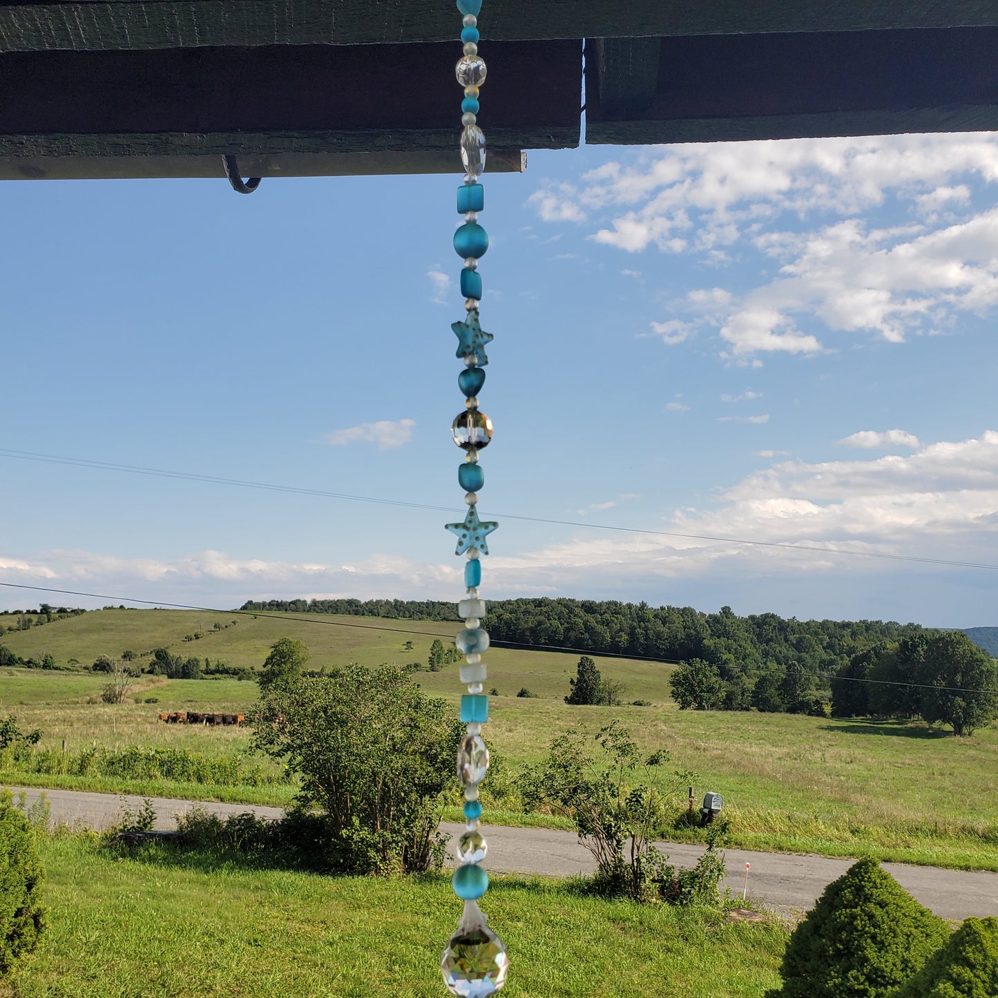 A beaded suncatcher hangs from a wooden eave, featuring turquoise, white, and clear glass beads with star shapes, set against a scenic rural background of green fields and a blue sky.