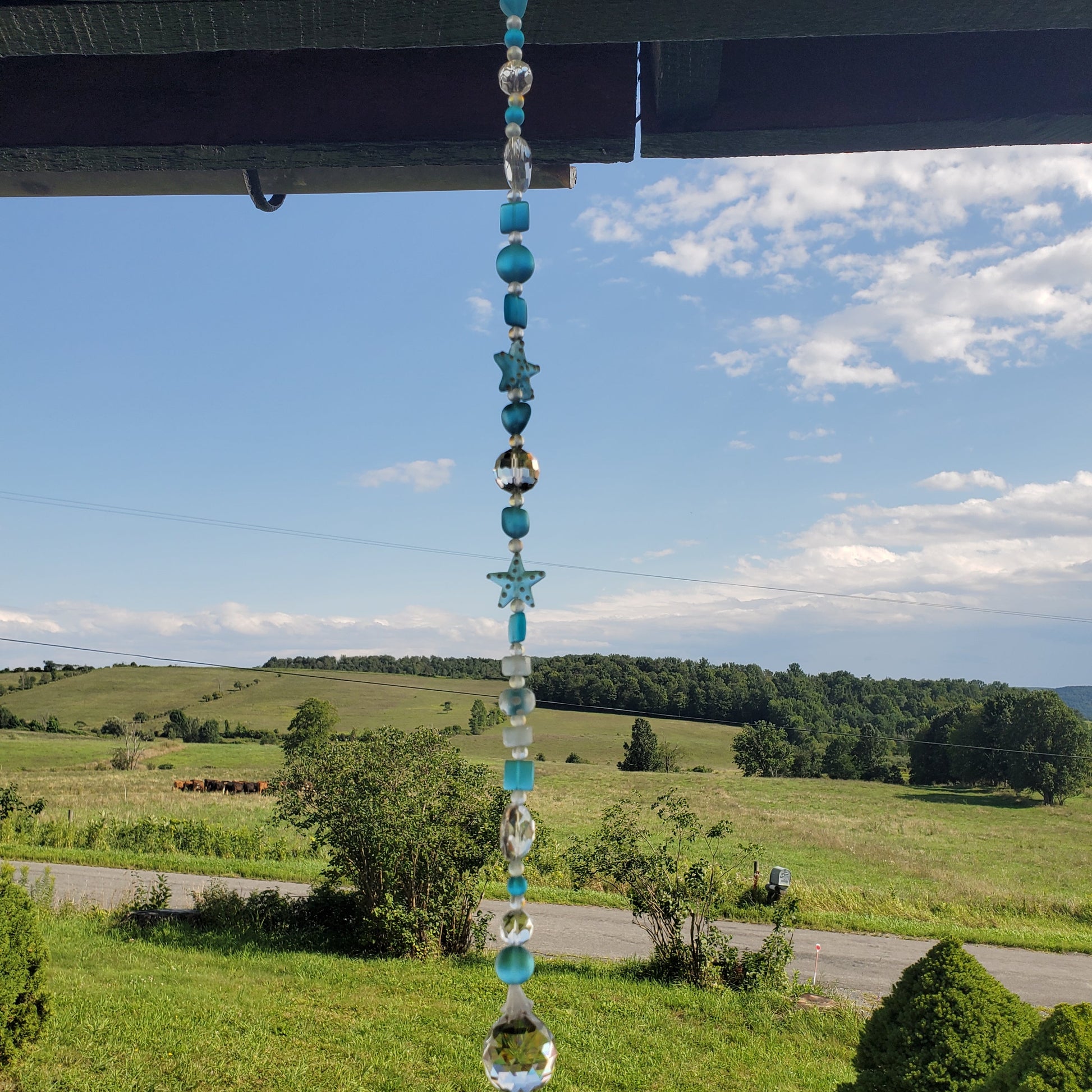 A beaded suncatcher hangs from a wooden eave, featuring turquoise, white, and clear glass beads with star shapes, set against a scenic rural background of green fields and a blue sky.