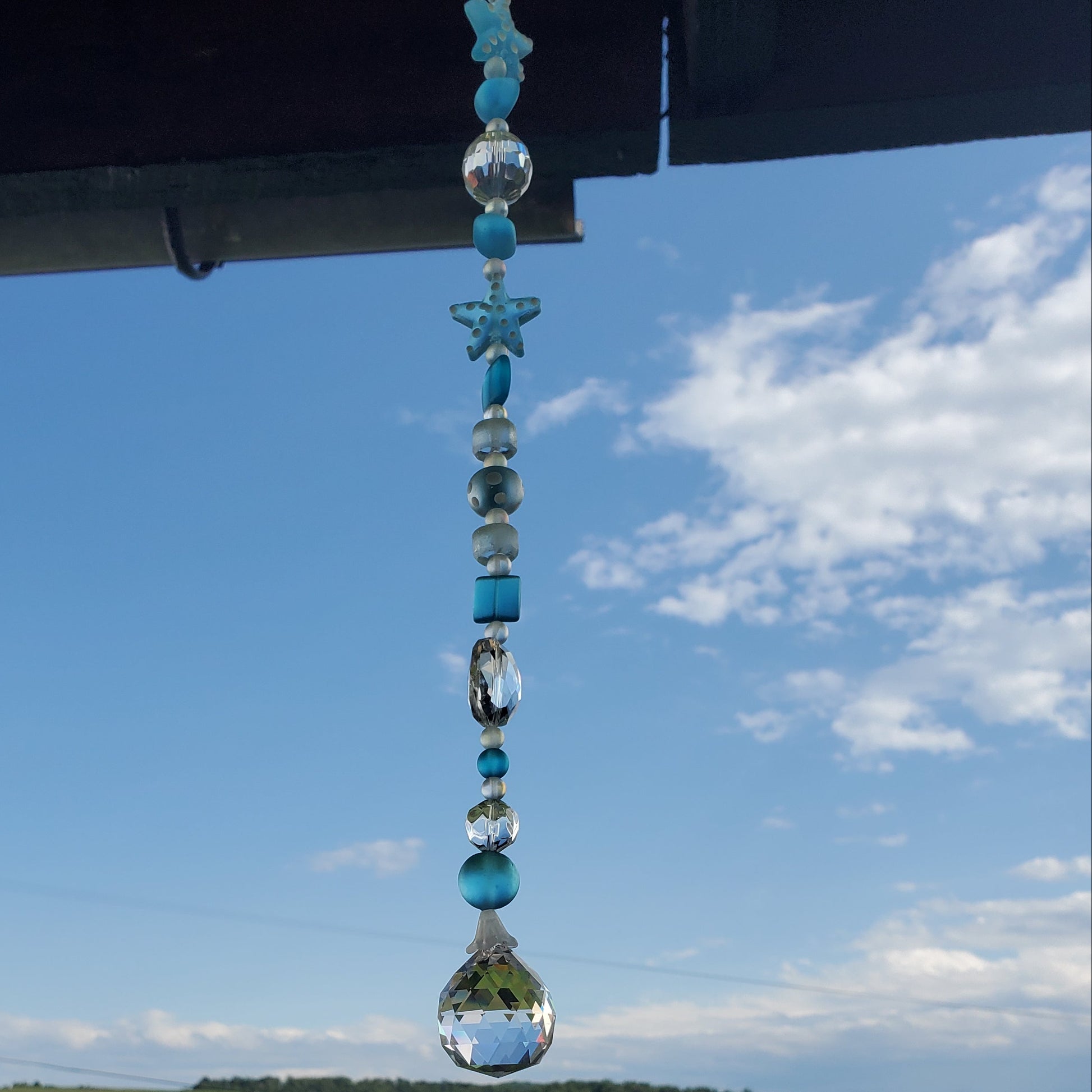 A hanging beaded suncatcher with turquoise blue and clear faceted beads, including a star and teardrop shapes, suspended under a roof overhang with a scenic green field and blue sky in the background.