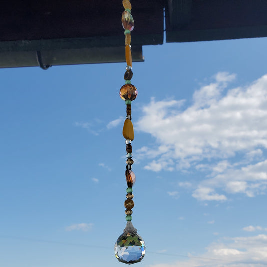 A hanging glass suncatcher ornament with amber and clear beads hangs outdoors under a roof eave, reflecting the blue sky and green fields below.