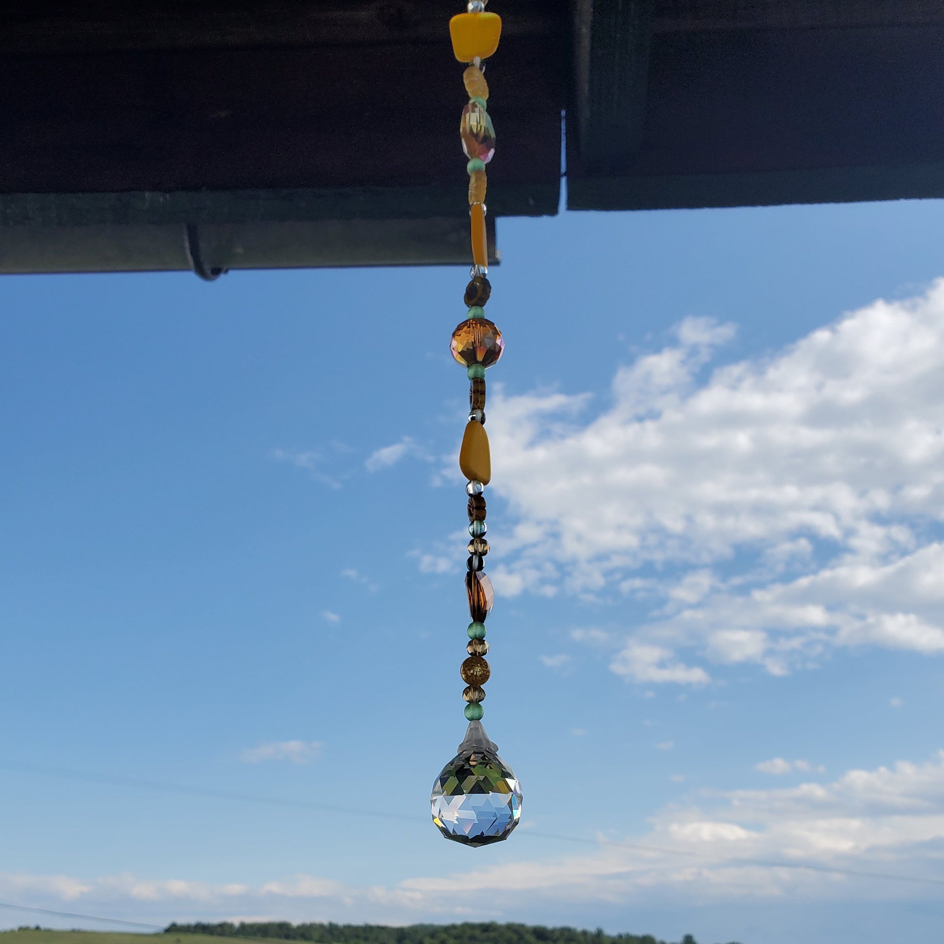 A hanging glass suncatcher with colorful decorative beads and a clear teardrop-shaped bottom, photographed outdoors under a roof overhang with a grassy field and blue sky in the background.