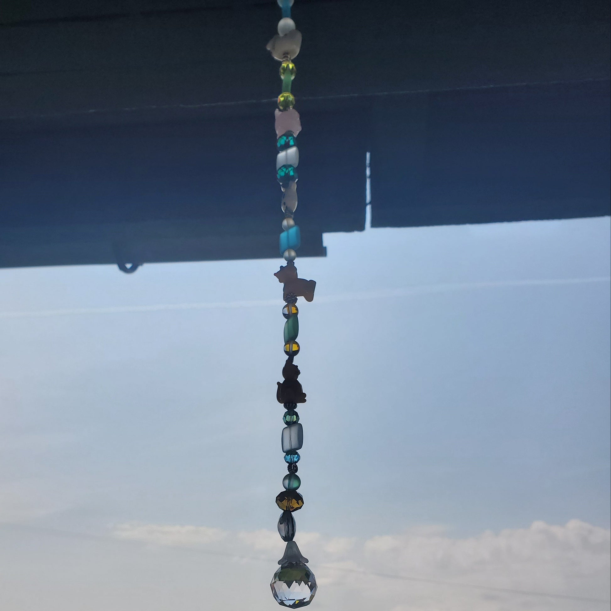 A beaded window suncatcher with colorful glass beads and a clear crystal raindrop hangs in front of a window overlooking a green rural landscape.