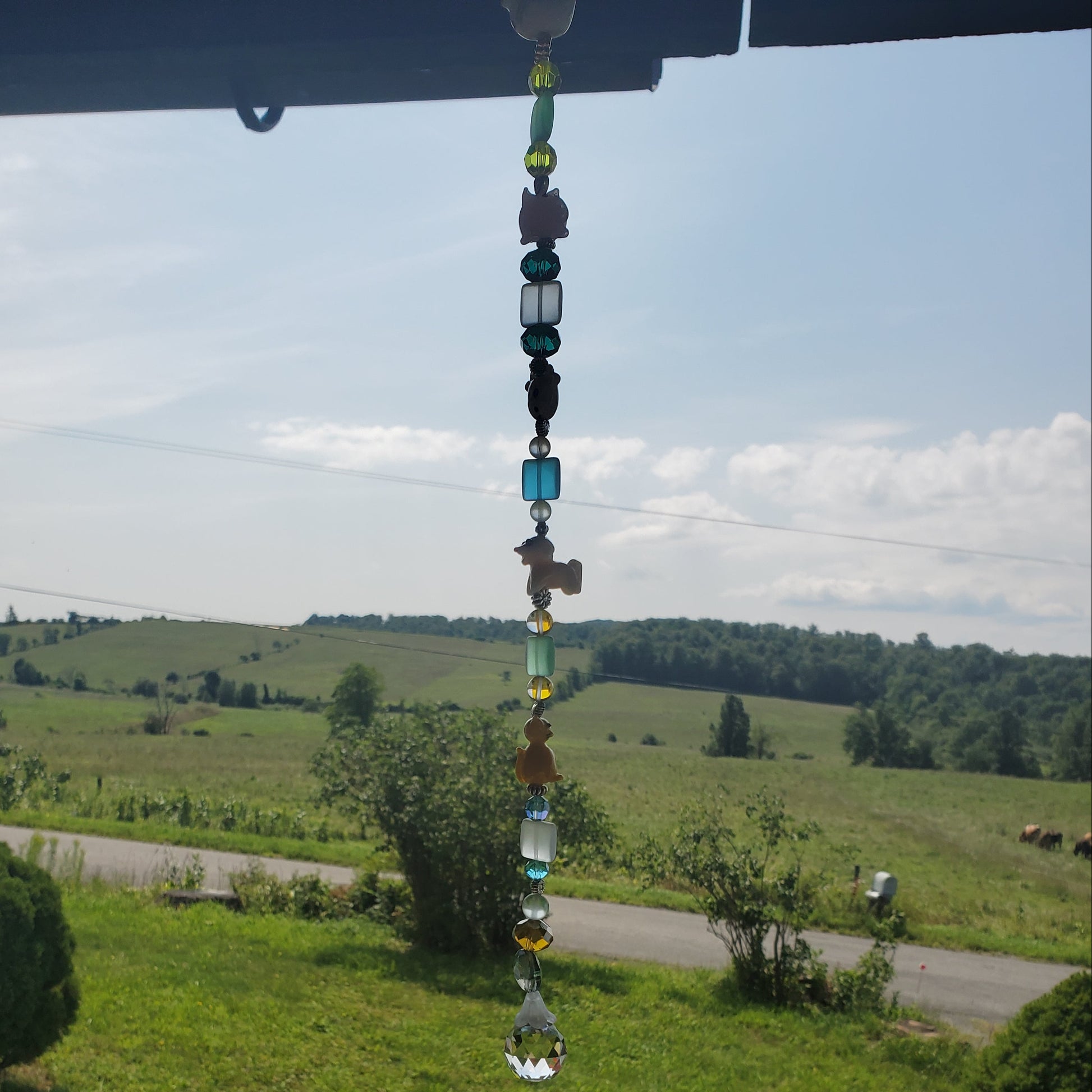 A beaded suncatcher with colorful glass beads hangs from a porch overhang, overlooking a green grassy yard and a rural road under a cloudy sky.