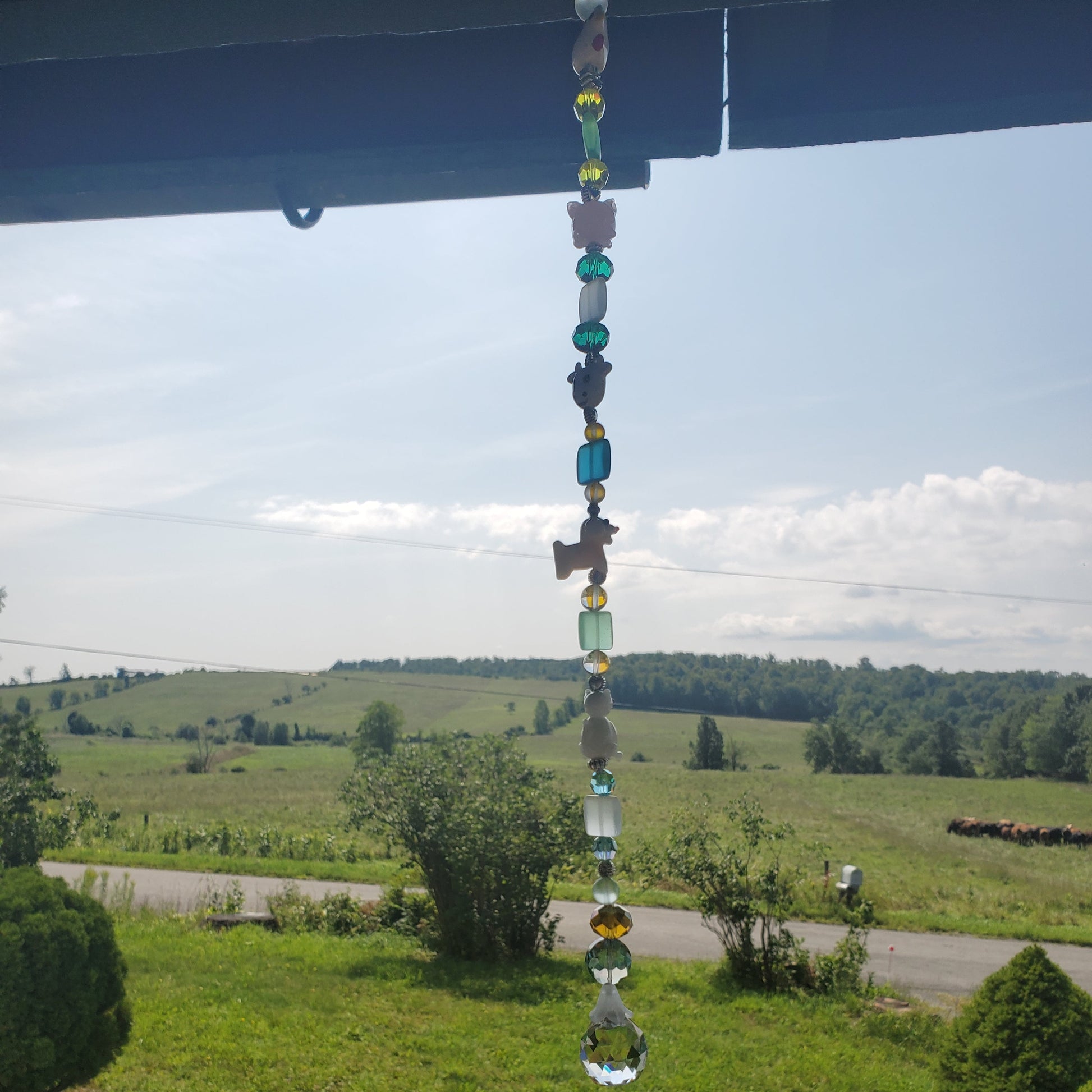A beaded suncatcher hangs from a roof overhang, with a rural landscape of green fields, trees, and a road visible in the background under a cloudy sky.
