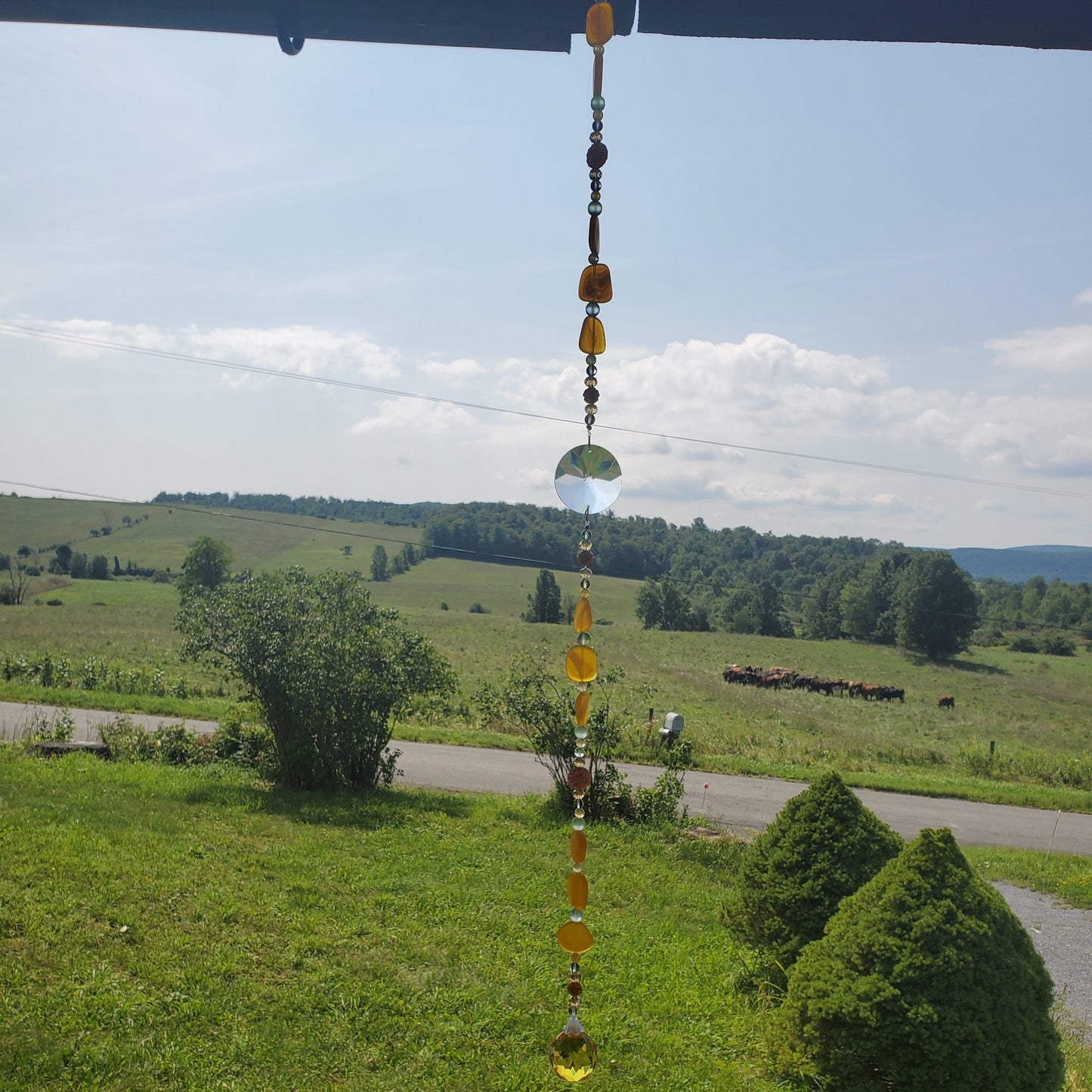 A hanging decorative suncatcher with amber and clear glass beads suspended outdoors in a grassy landscape with trees and a road under a blue sky.
