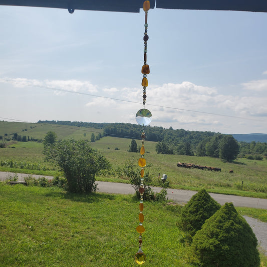 A hanging decorative suncatcher with amber and clear glass beads suspended outdoors in a grassy landscape with trees and a road under a blue sky.