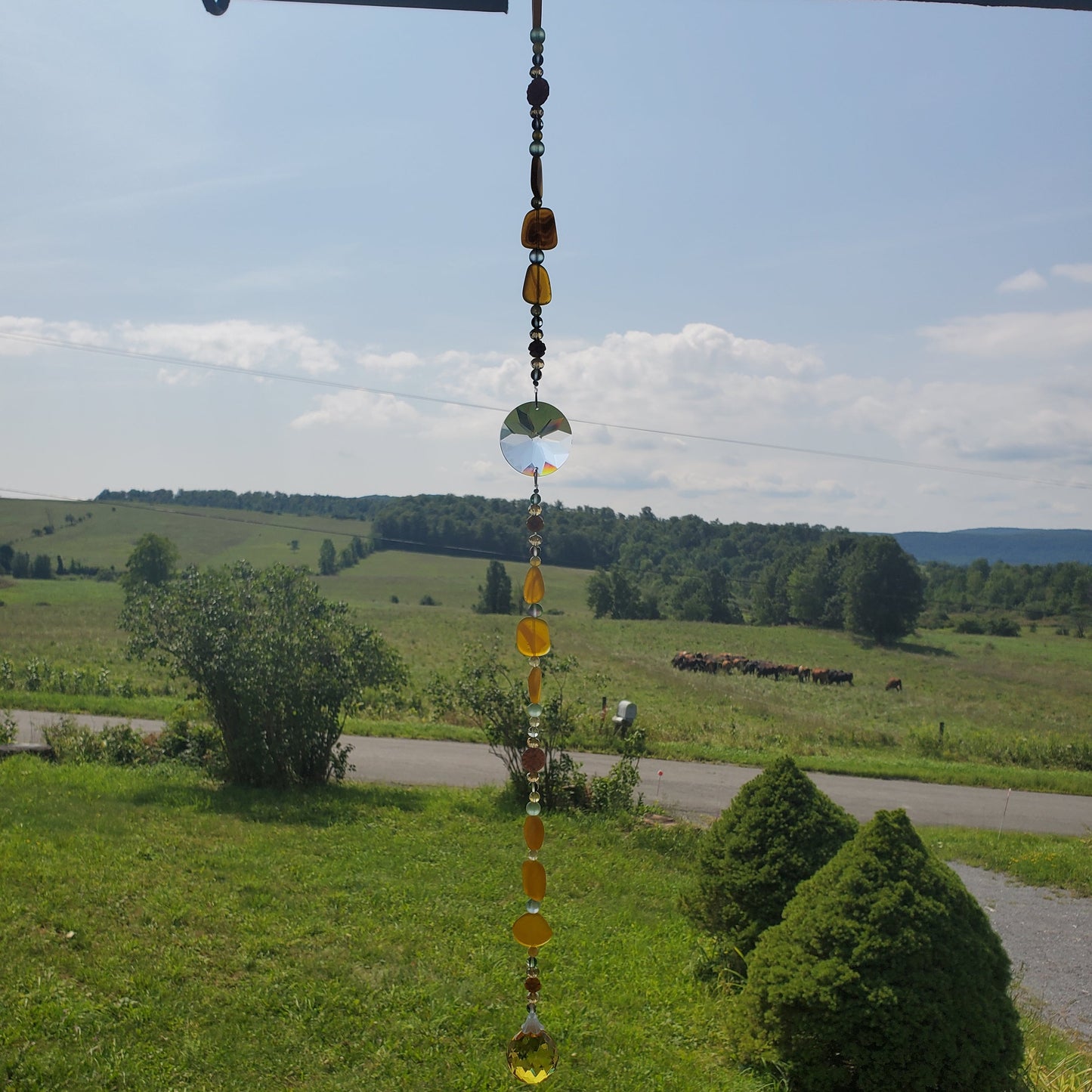 A hanging glass suncatcher with amber and clear beads is suspended outdoors in front of a scenic rural landscape with green fields, trees, and a road under a blue sky.