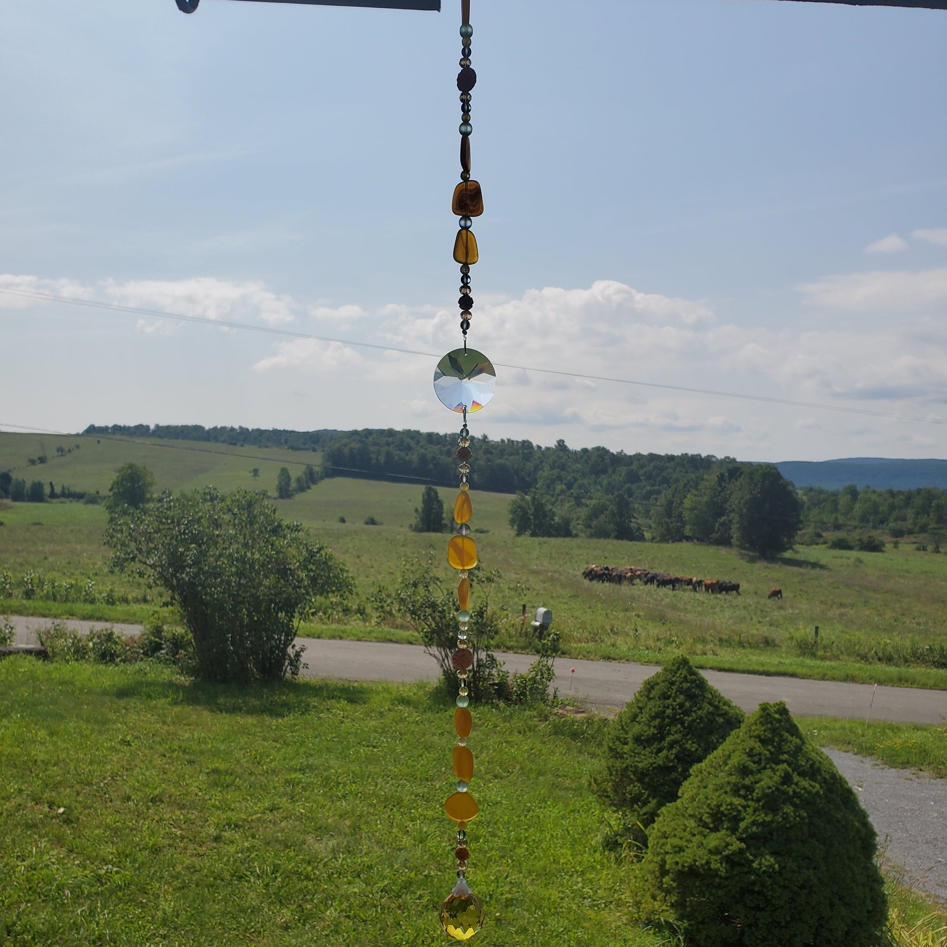 A hanging glass suncatcher with amber and clear beads is suspended outdoors in front of a scenic rural landscape with green fields, trees, and a road under a blue sky.