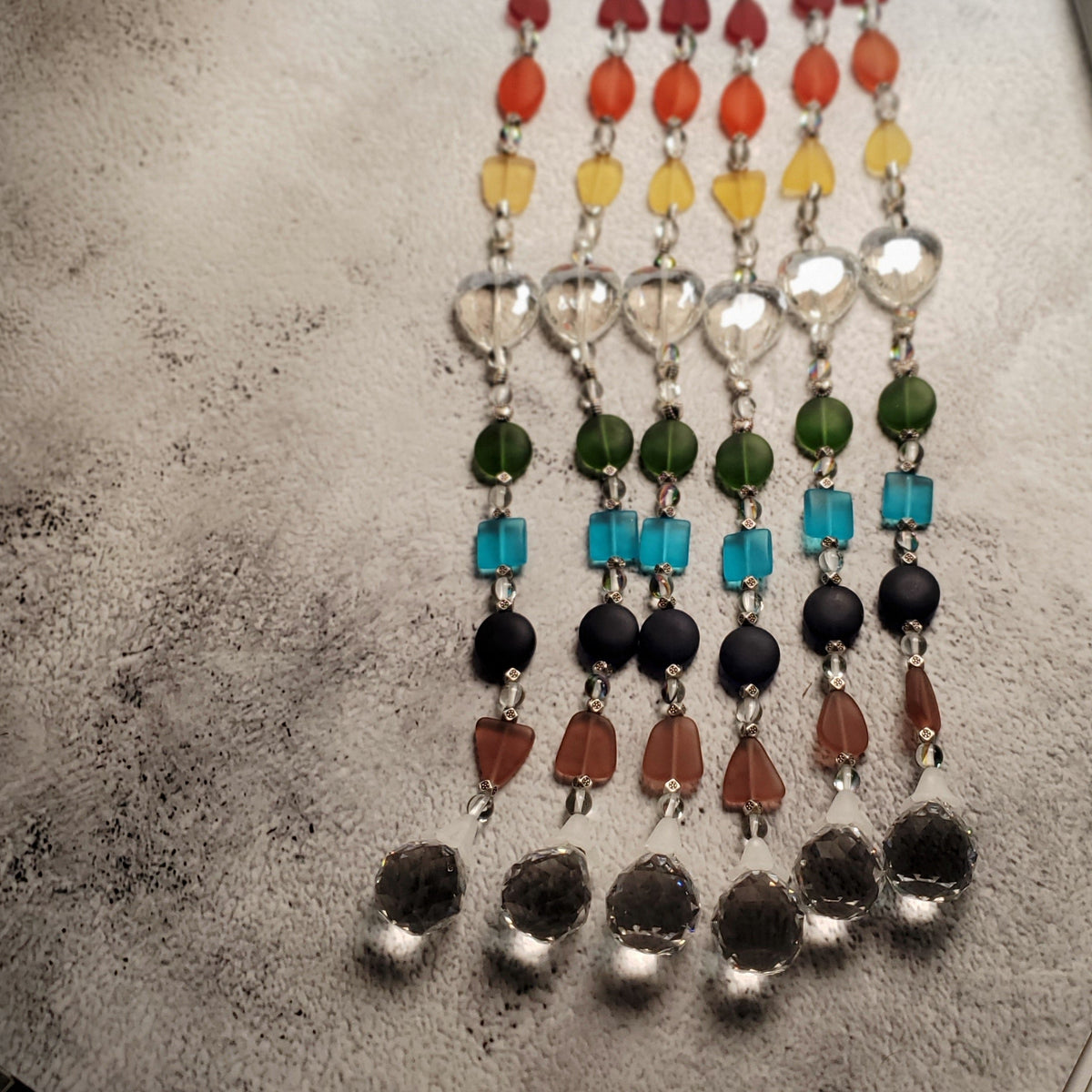 Six beaded rainbow suncatchers arranged vertically on a textured gray surface, featuring multicolored beads and crystal beads.