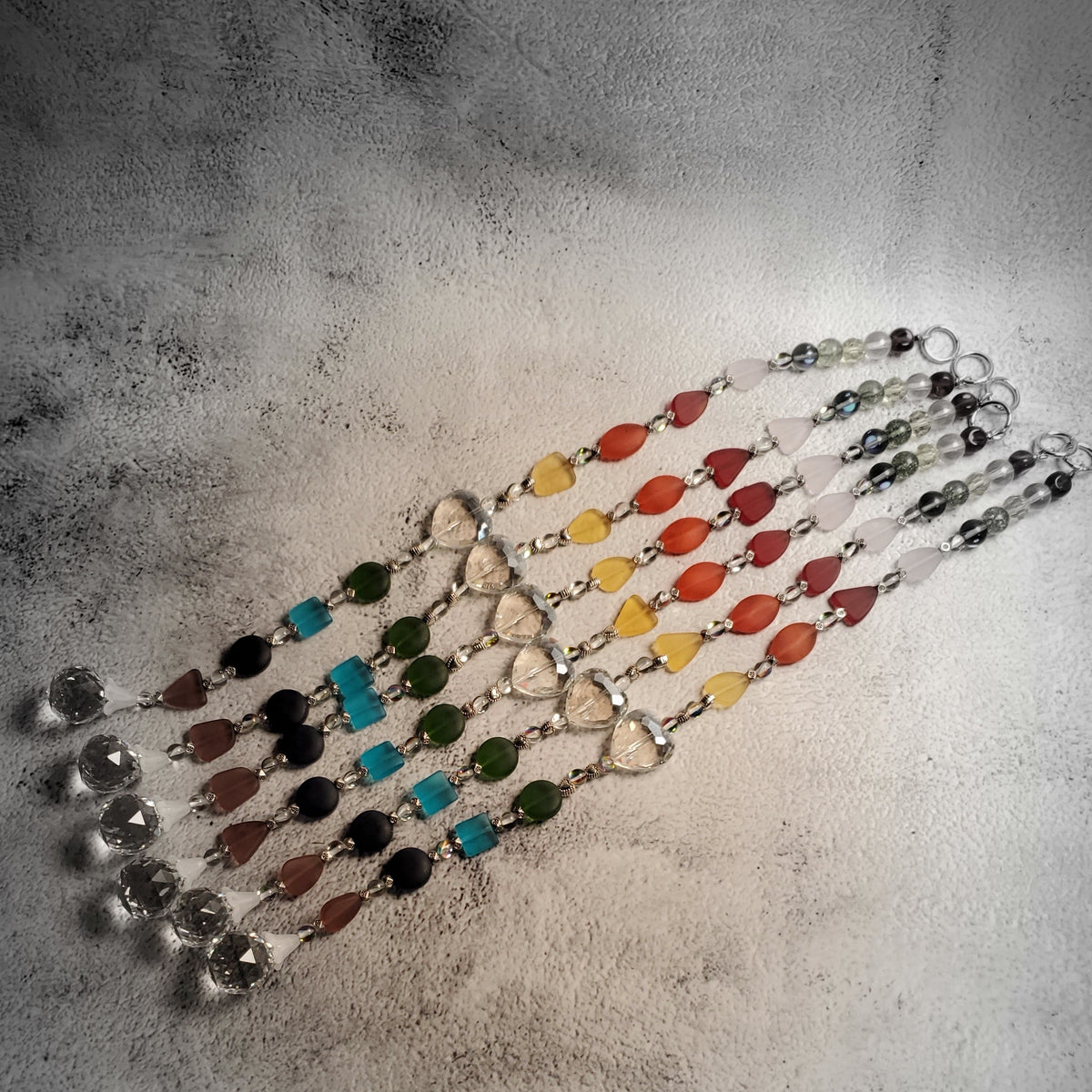 Beaded colorful suncatchers laid diagonally on a textured gray surface. Suncatchers feature clear faceted beads, and colored sea glass beads representing a rainbow sequence.