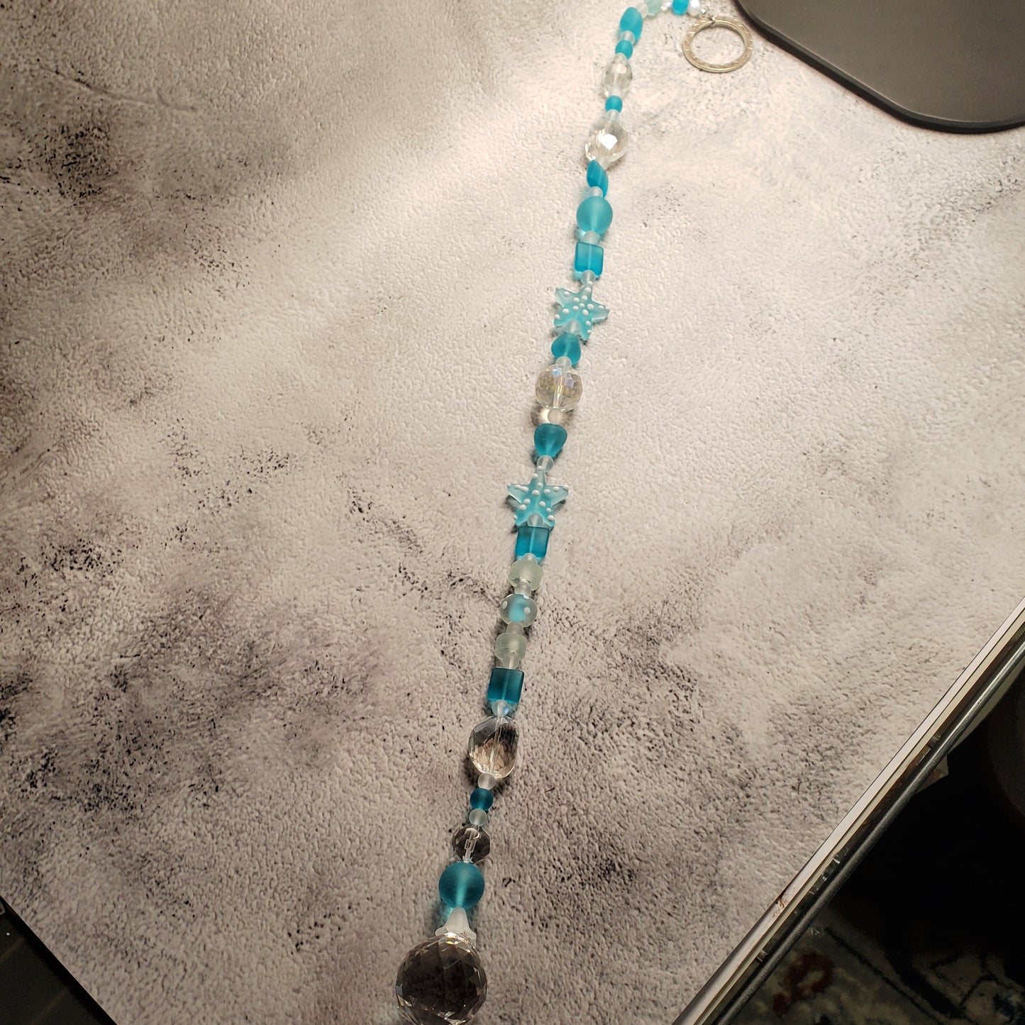 A beaded suncatcher laid diagonally on a textured gray surface, featuring turquoise blue beads, clear faceted beads, and a silver-toned ring.