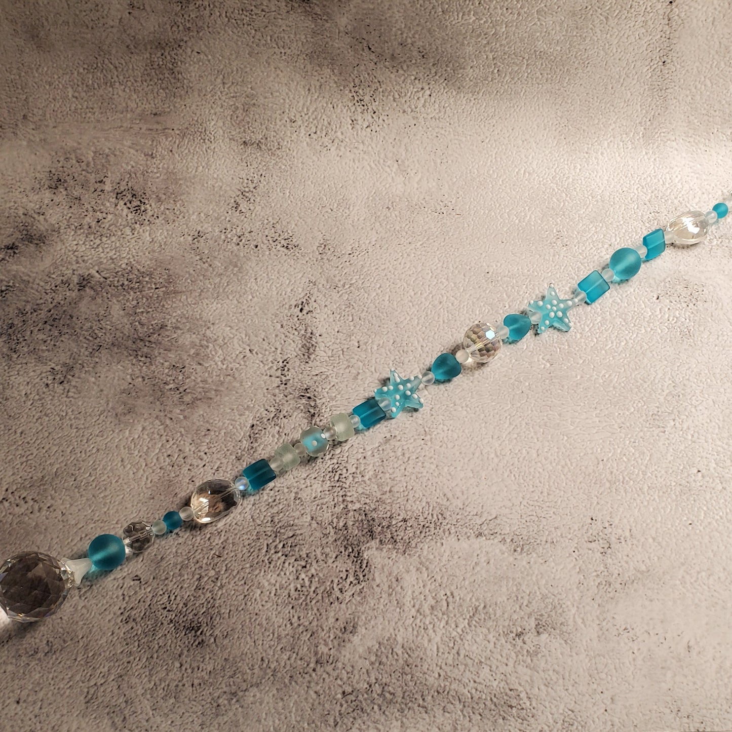 A beaded suncatcher with turquoise blue, light blue, and silver beads draped diagonally across a textured gray backdrop.