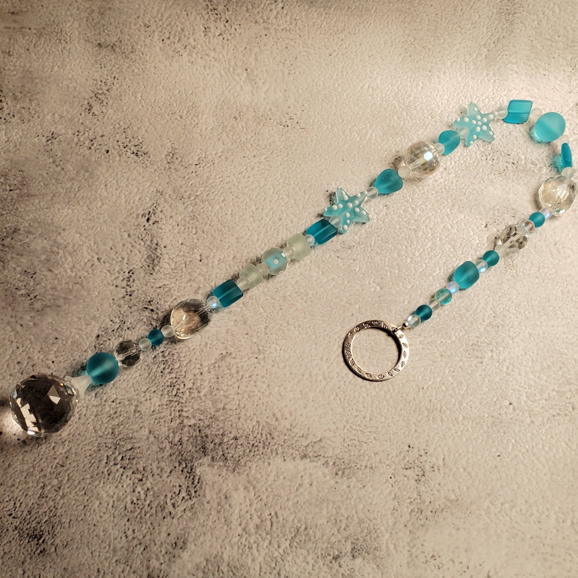 A beaded suncatcher with turquoise blue, clear, and white beads, featuring a silver-toned metal O-ring at one end, displayed on a textured concrete surface.