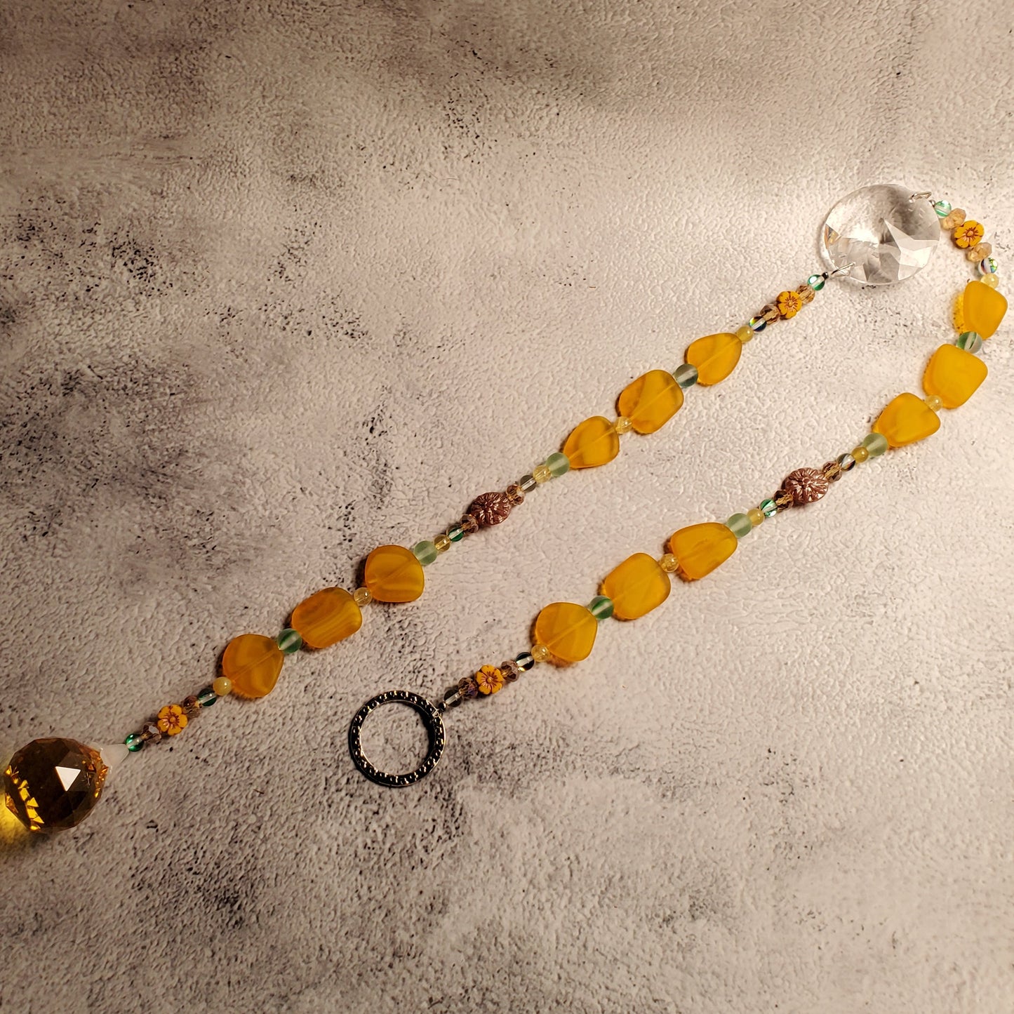 A beaded suncatcher with translucent yellow-orange beads, small yellow spacer beads, and clear faceted crystal end piece, laid on a textured light gray surface.