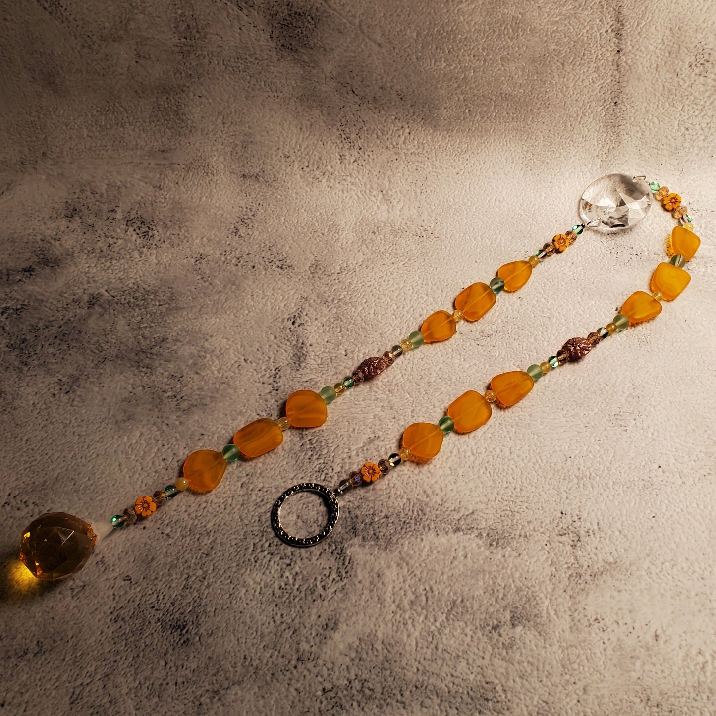 A beaded suncatcher with orange and yellow sea glass beads, green accents, and a clear faceted crystal, displayed on a textured gray surface.